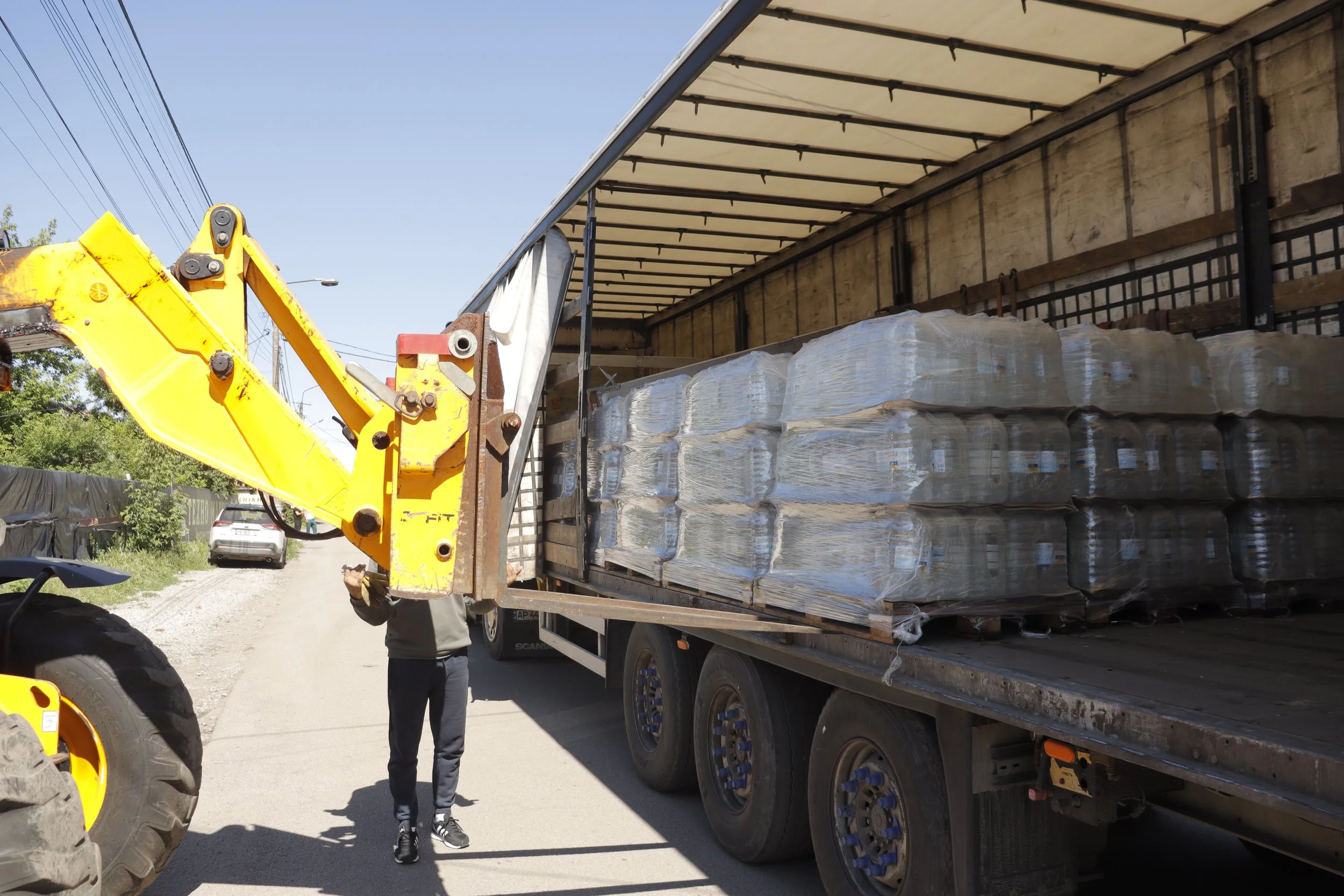 A person standing next to a large truck filled with pallets wrapped in plastic, with a yellow construction vehicle arm touching the truck.