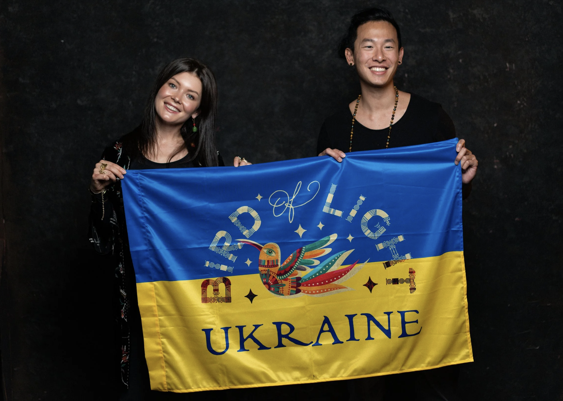 Two smiling people, one woman and one man, holding a Ukrainian flag with a colorful bird design and the words "of Light" and "Ukraine" on it. They are standing against a dark background.
