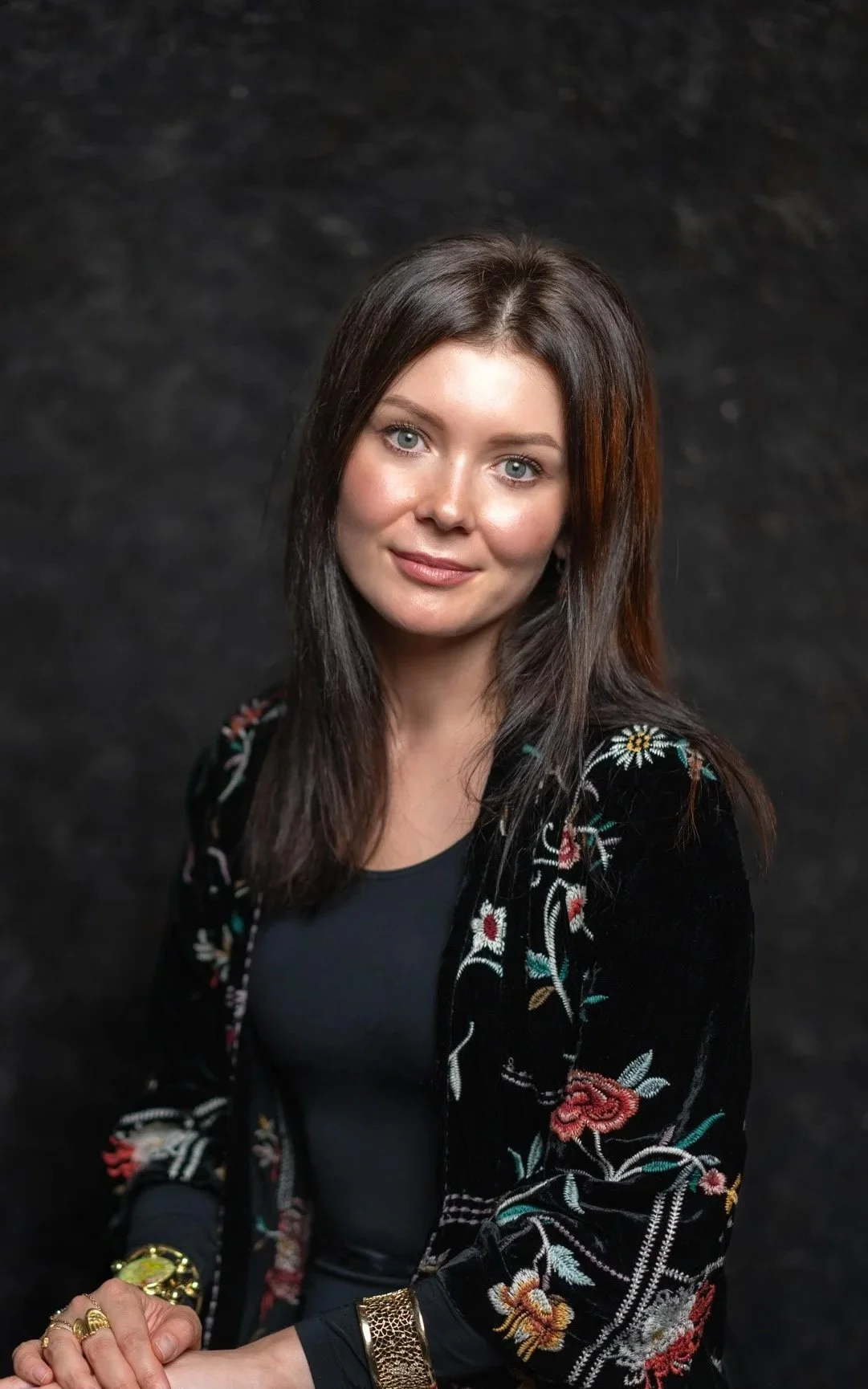 A woman with long dark hair, wearing a black embroidered jacket and gold jewelry, posing against a dark background.