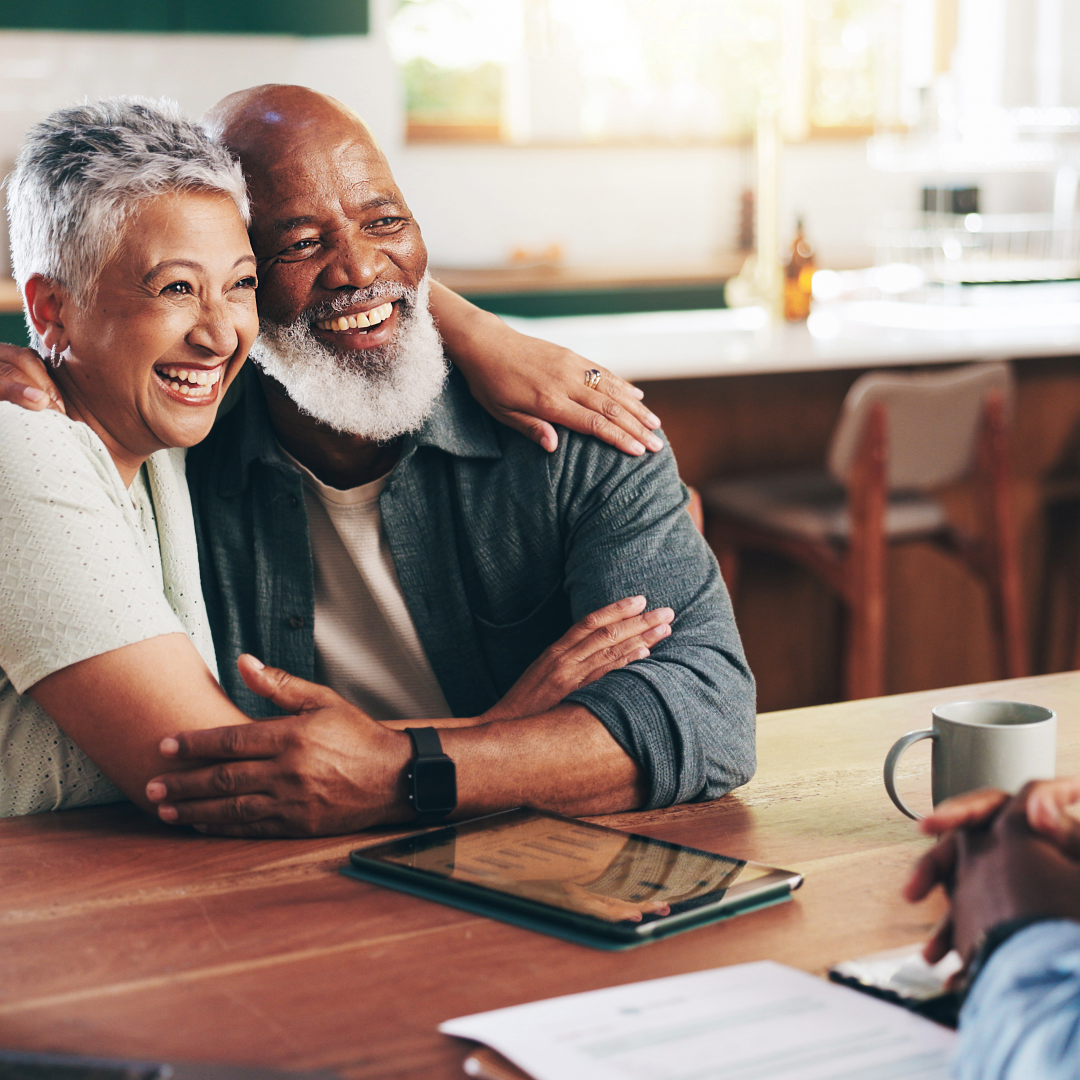 An elderly couple sitting at a table, smiling and hugging each other warmly, preparing for debt free life & retirement!