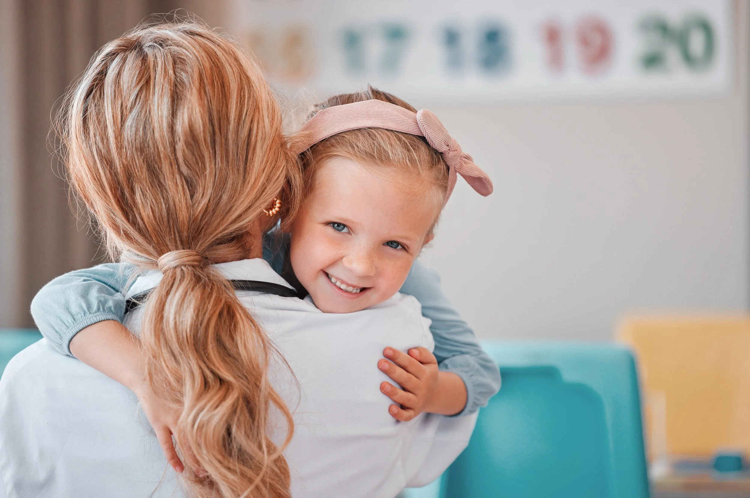 A smiling young girl with a pink headband hugging a speech therapist.
