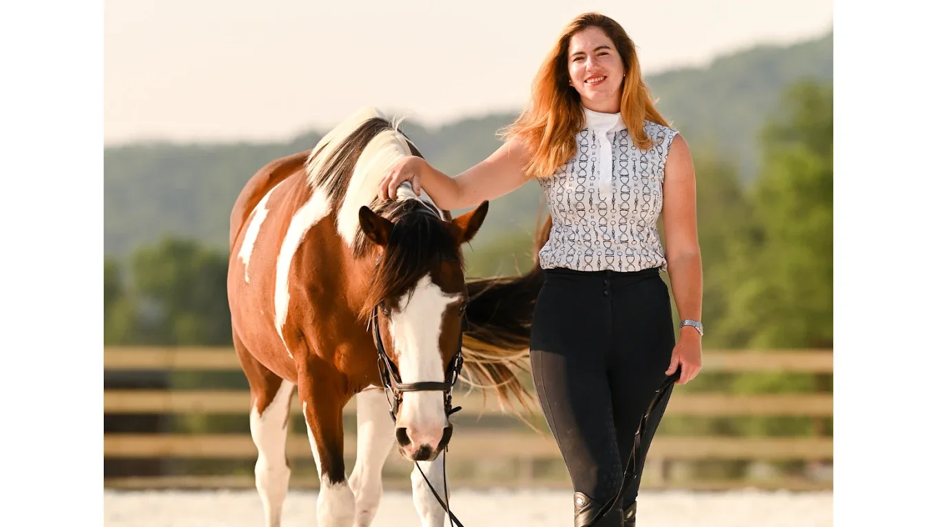 A woman with long reddish-brown hair smiling and holding the reins of a brown and white pinto horse outdoors with a blurred green background.