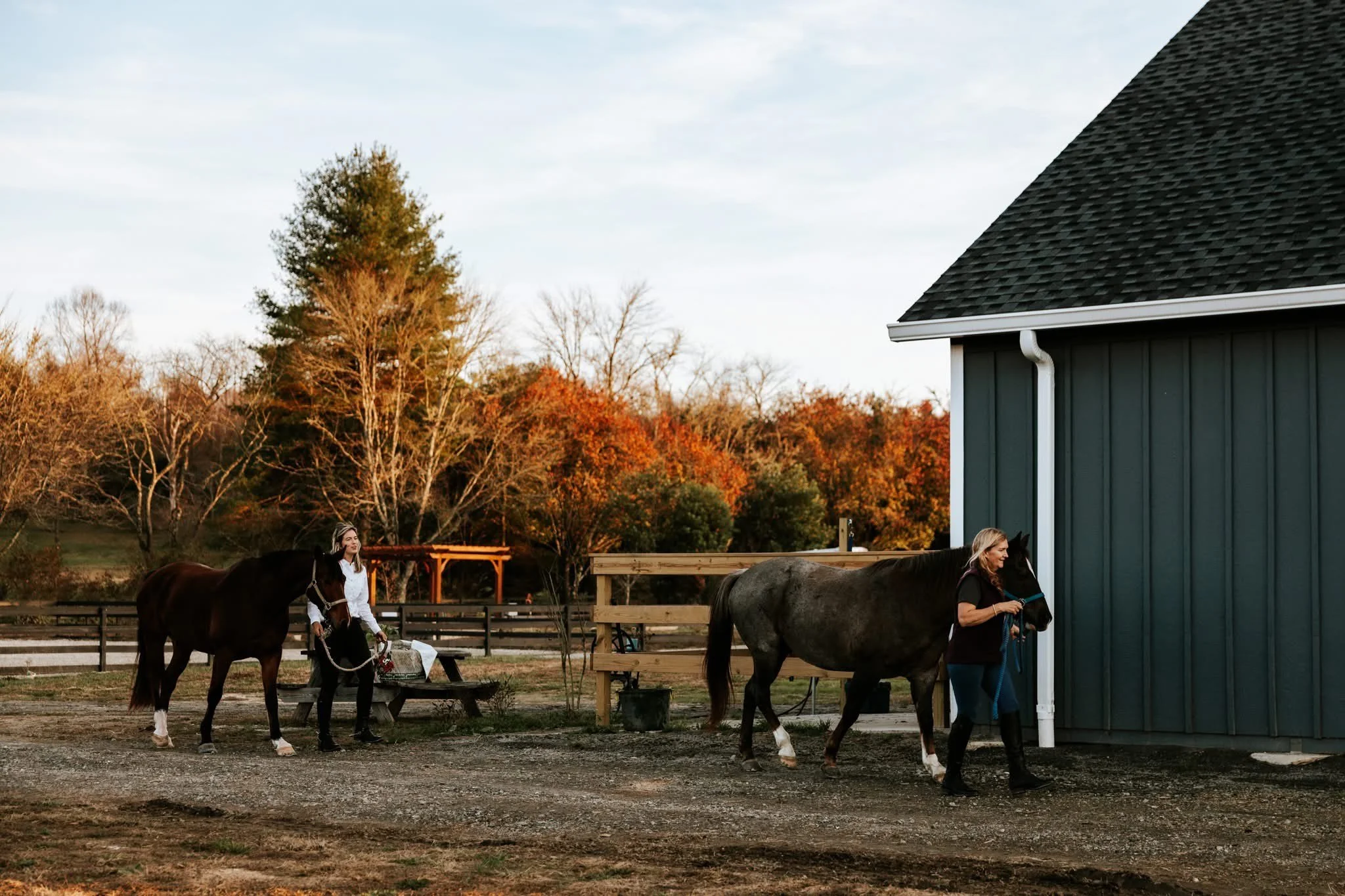 Two women with horses outside a green barn during autumn, with trees in fall foliage in the background.