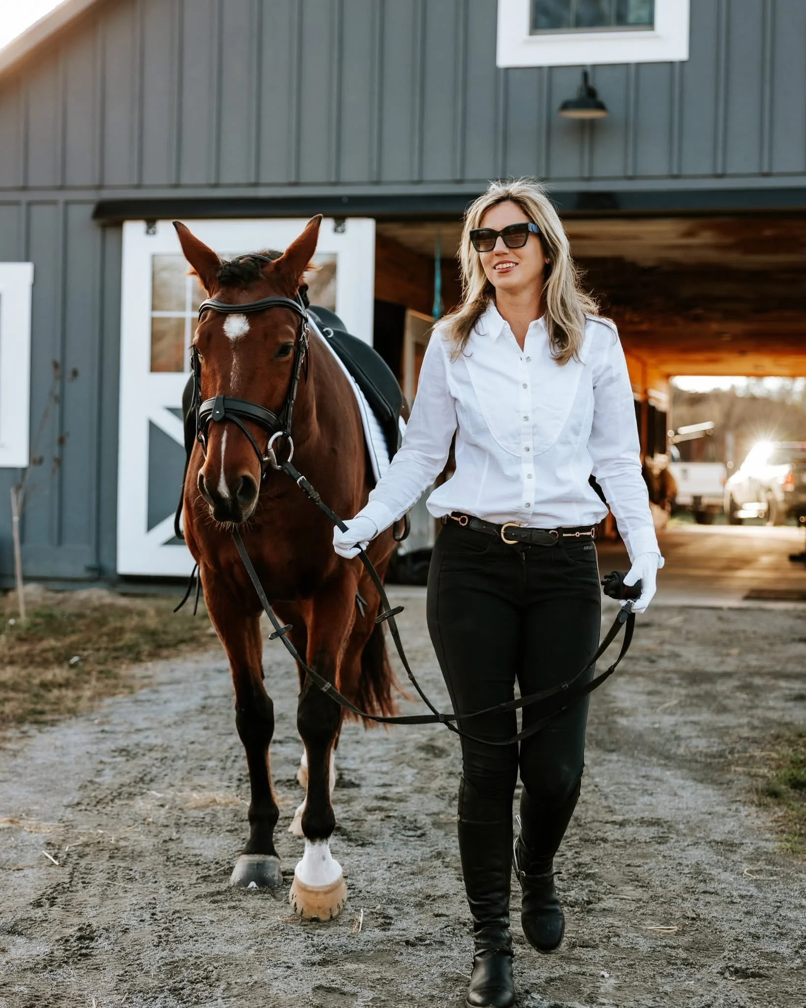 A woman walking a brown horse outside a barn on a farm during sunset.