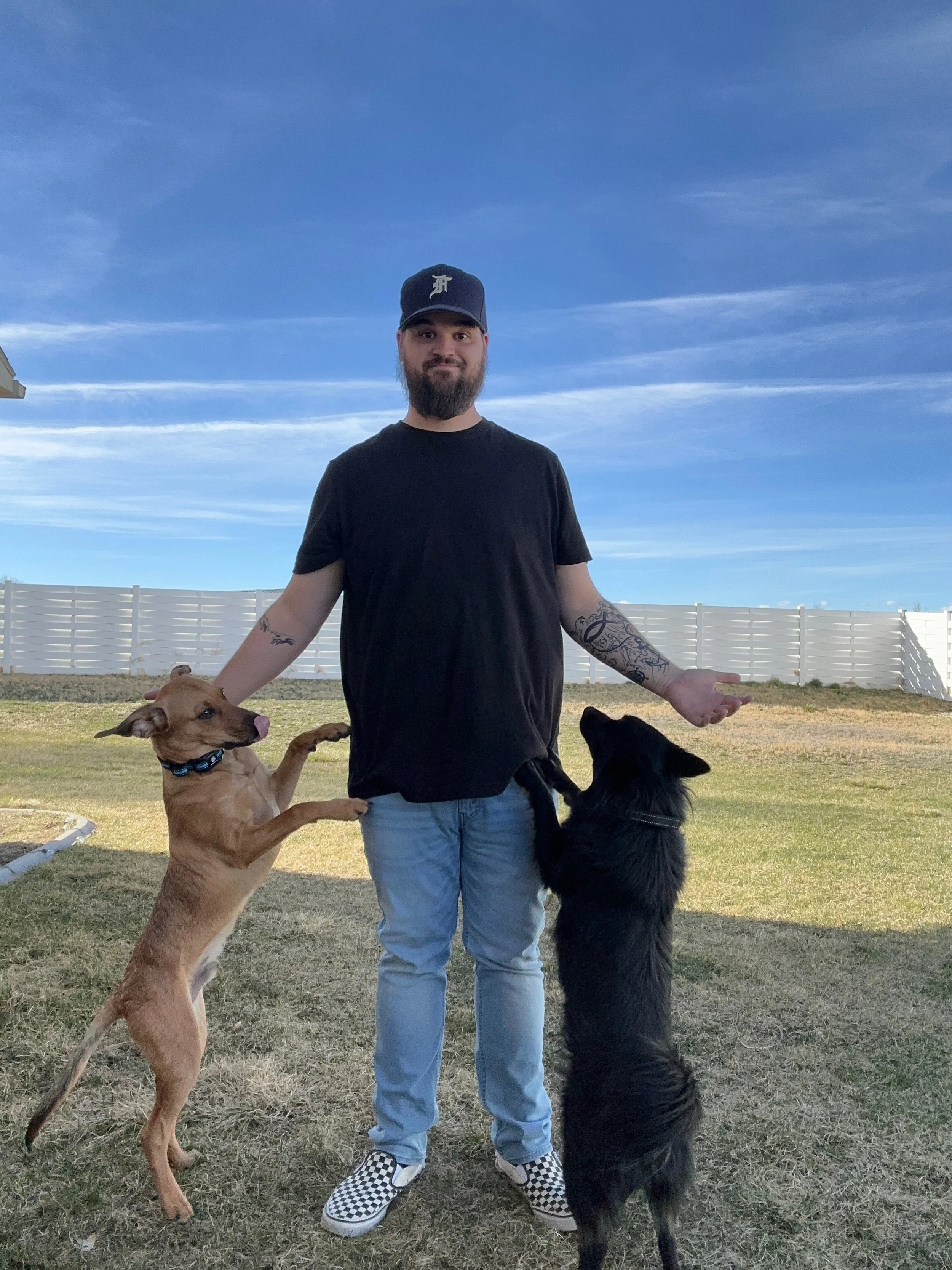 A man with a beard and tattoos, wearing a black T-shirt, jeans, checkered slip-on shoes, and a baseball cap, standing in a backyard with two dogs, one tan and one black, both standing on their hind legs with the front paws on his arms, under a blue sky with some clouds.