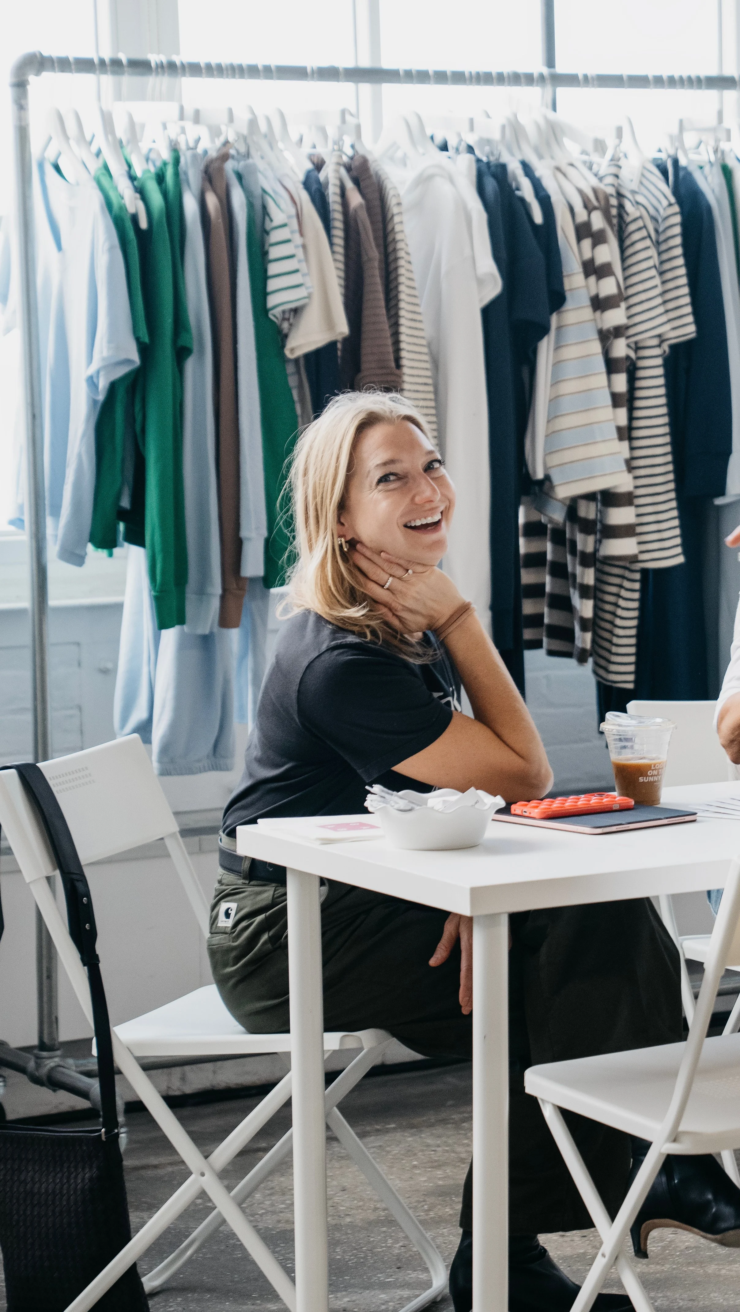 A woman smiling and sitting at a table in a room with clothing on racks behind her.