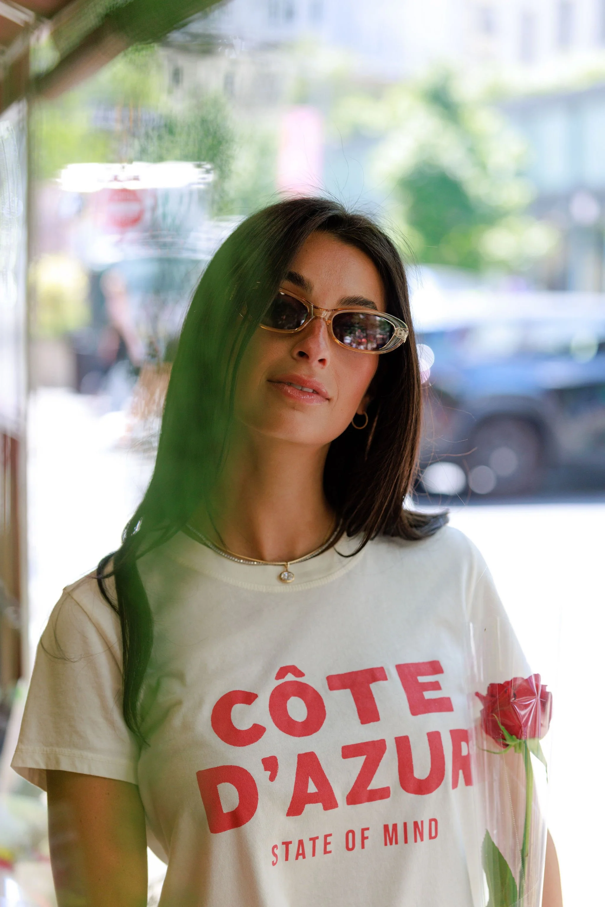 A woman wearing sunglasses and a white t-shirt with red text, standing near a store window with a blurred outdoor background, and a red flower tucked into her sleeve.