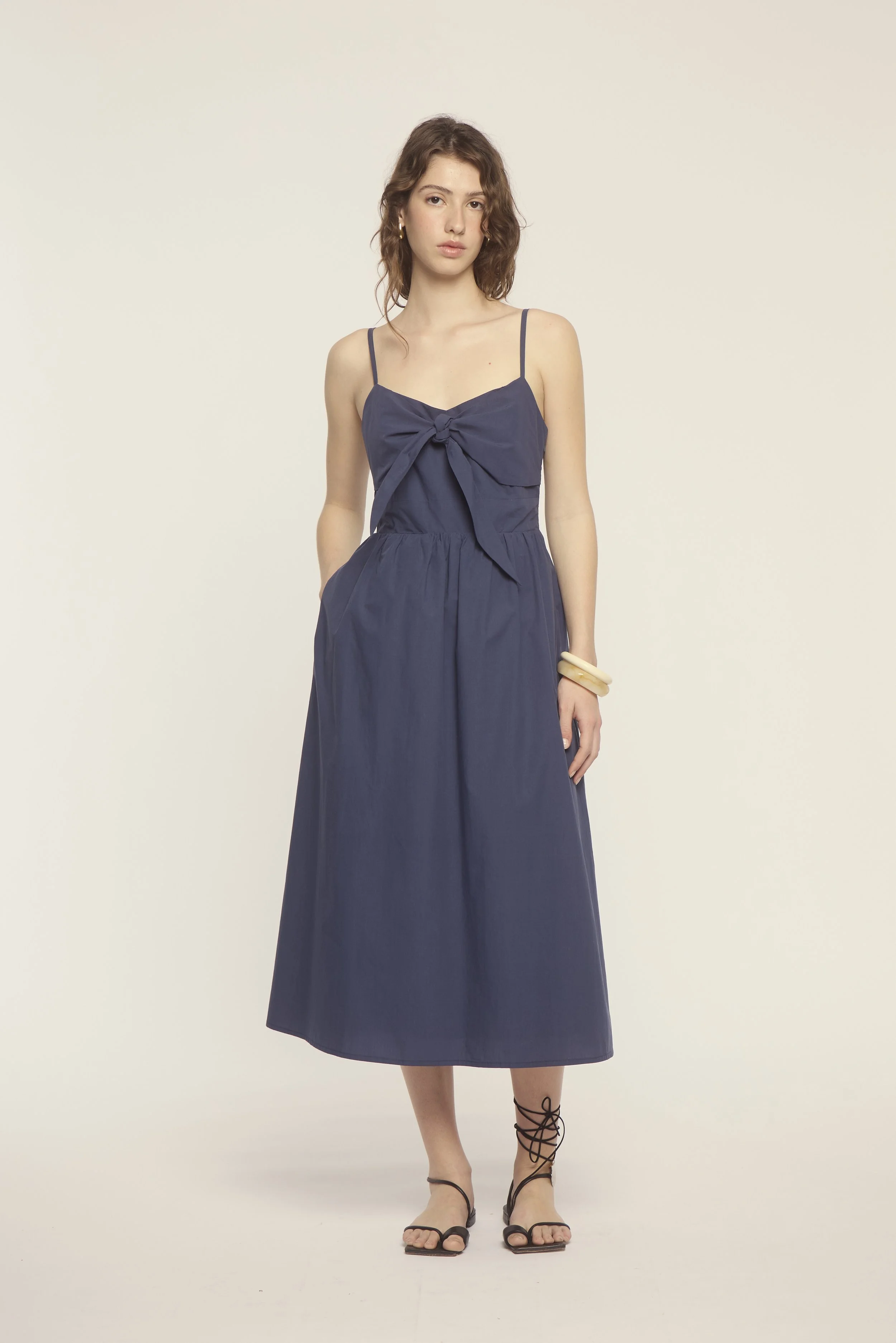 A young woman with wavy brown hair wearing a navy blue sleeveless midi dress with spaghetti straps and a front tie bow, standing against a plain light background.