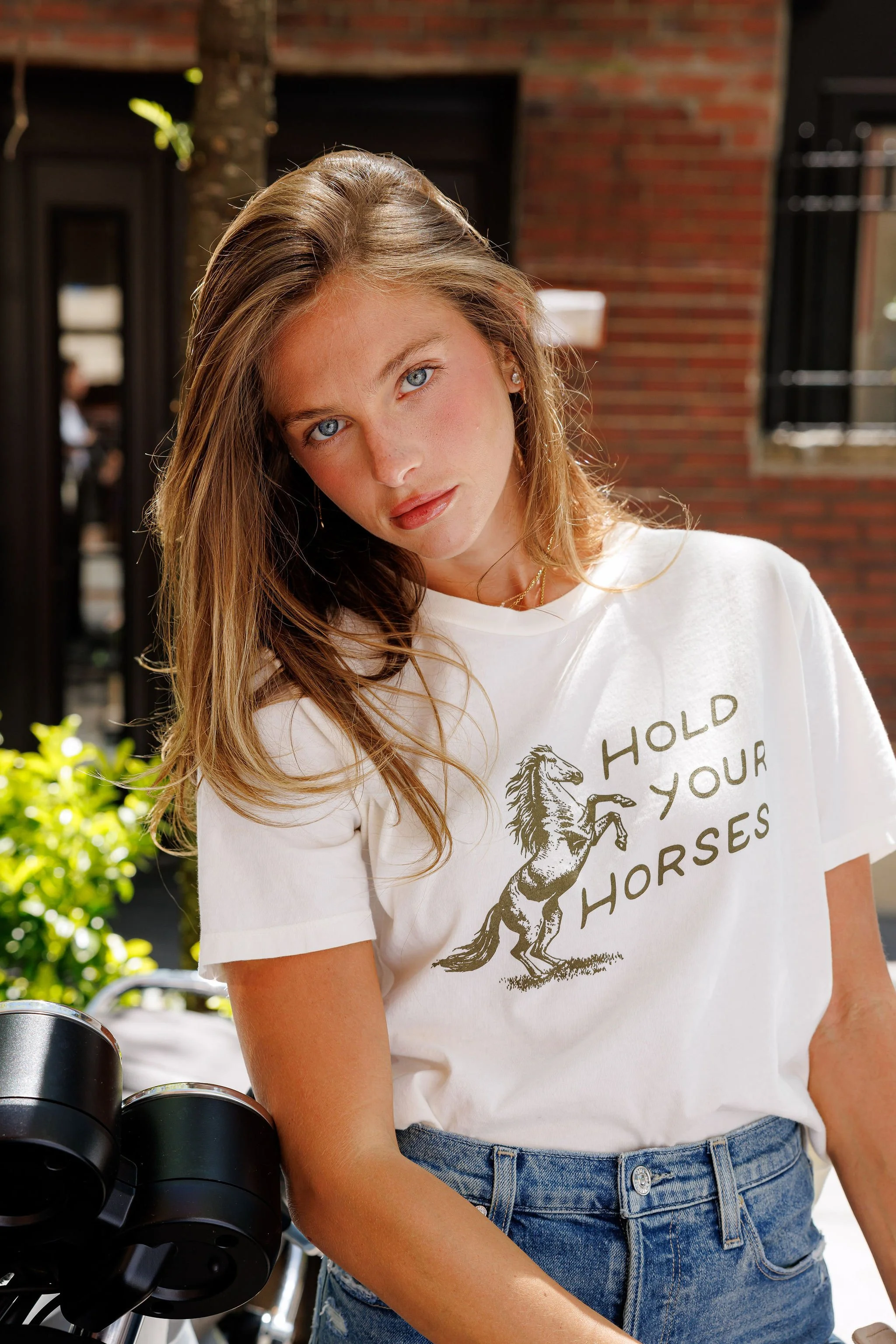 A young woman with long, light brown hair and blue eyes wearing a white t-shirt with a horse graphic and the words 'HOLD YOUR HORSES,' standing outdoors near a motorcycle, with a brick building and greenery in the background.
