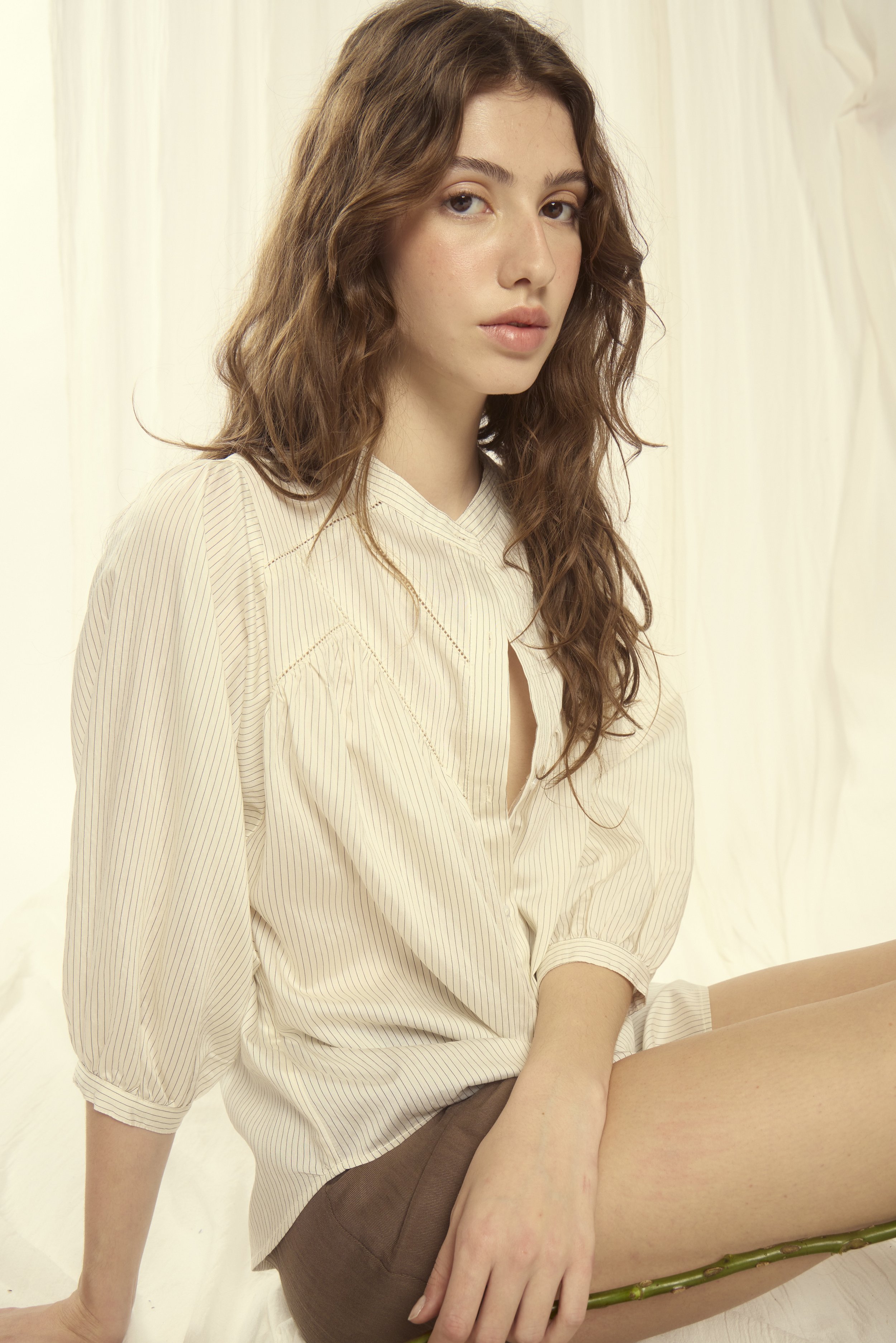 A young woman with wavy brown hair, wearing a beige striped blouse, is sitting against a light background.