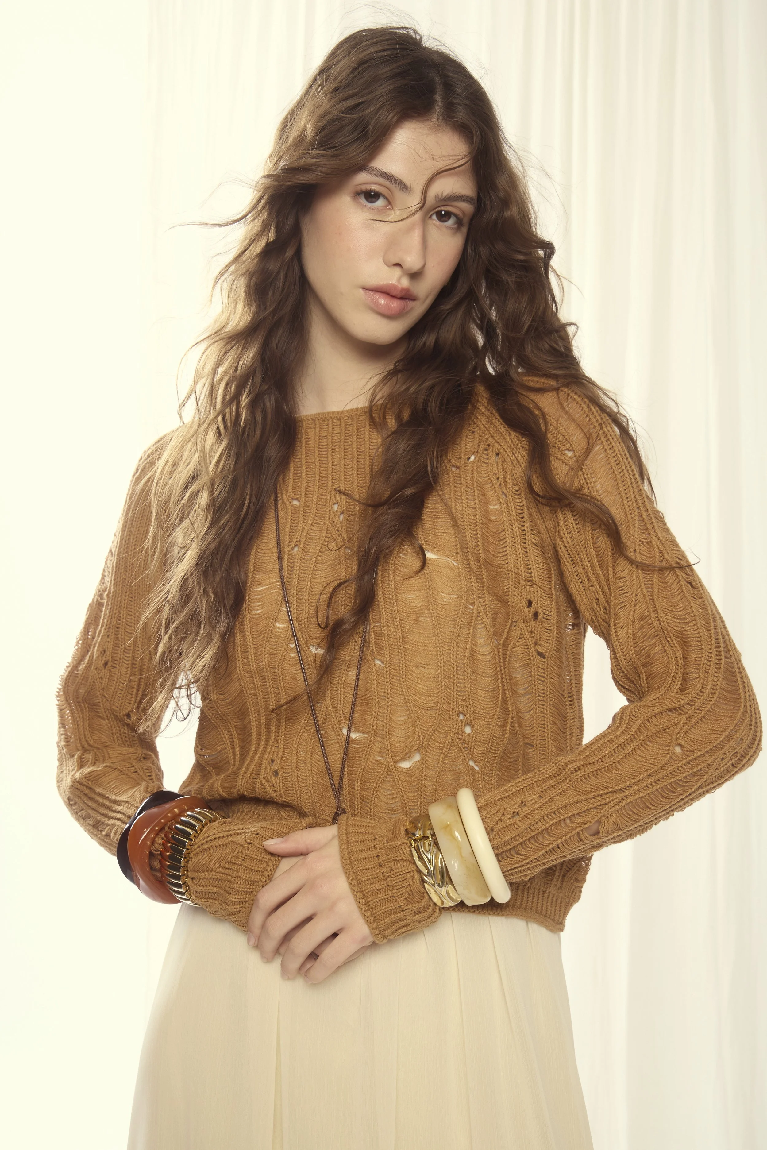 A young woman with long, wavy brown hair wearing a tan, distressed knit sweater and a cream skirt, standing indoors near sheer white curtains.