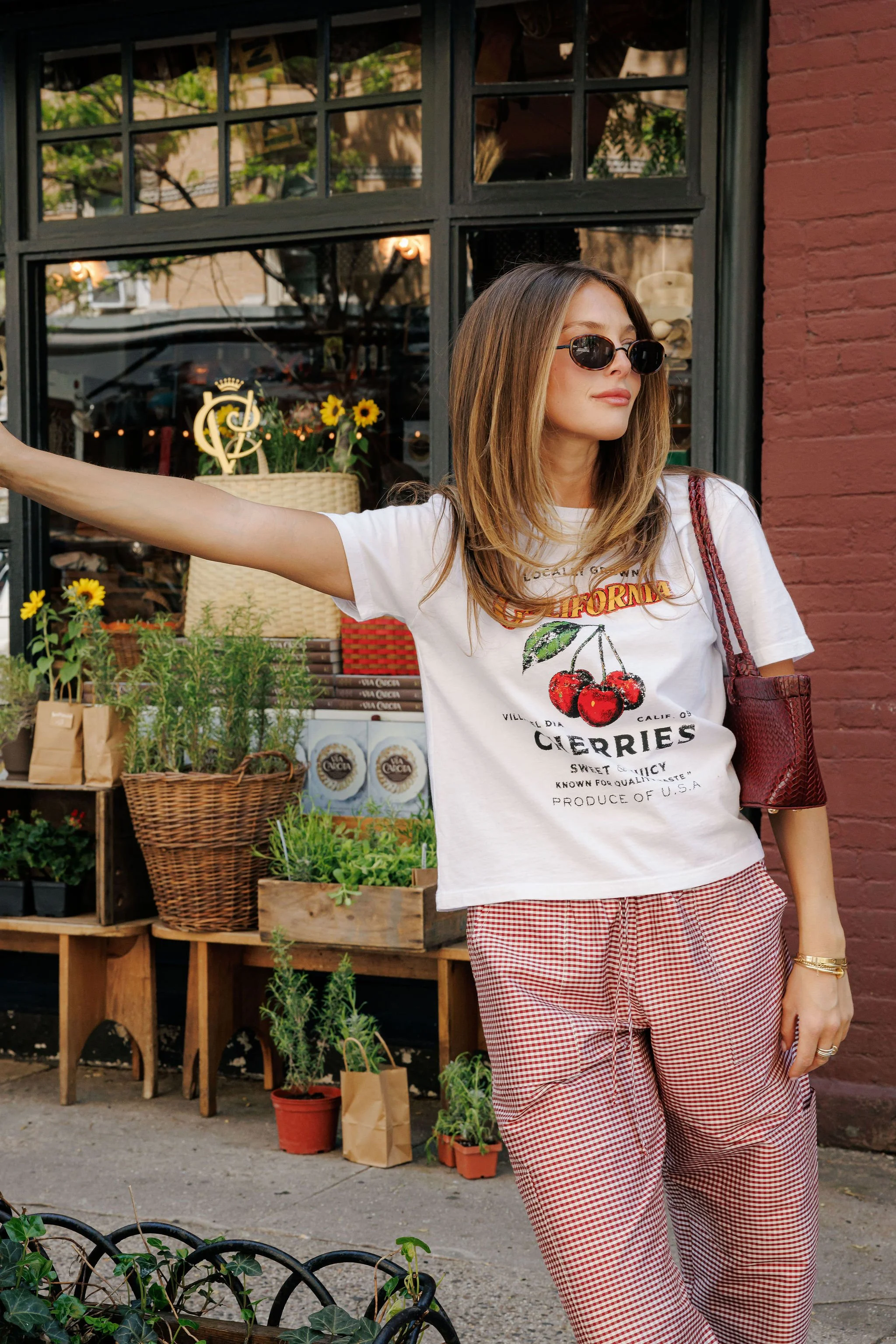A woman with shoulder-length brown hair wearing sunglasses, a white cherry graphic T-shirt, and red checkered pants stands outside a shop with potted plants and sunflowers, holding her arm out.