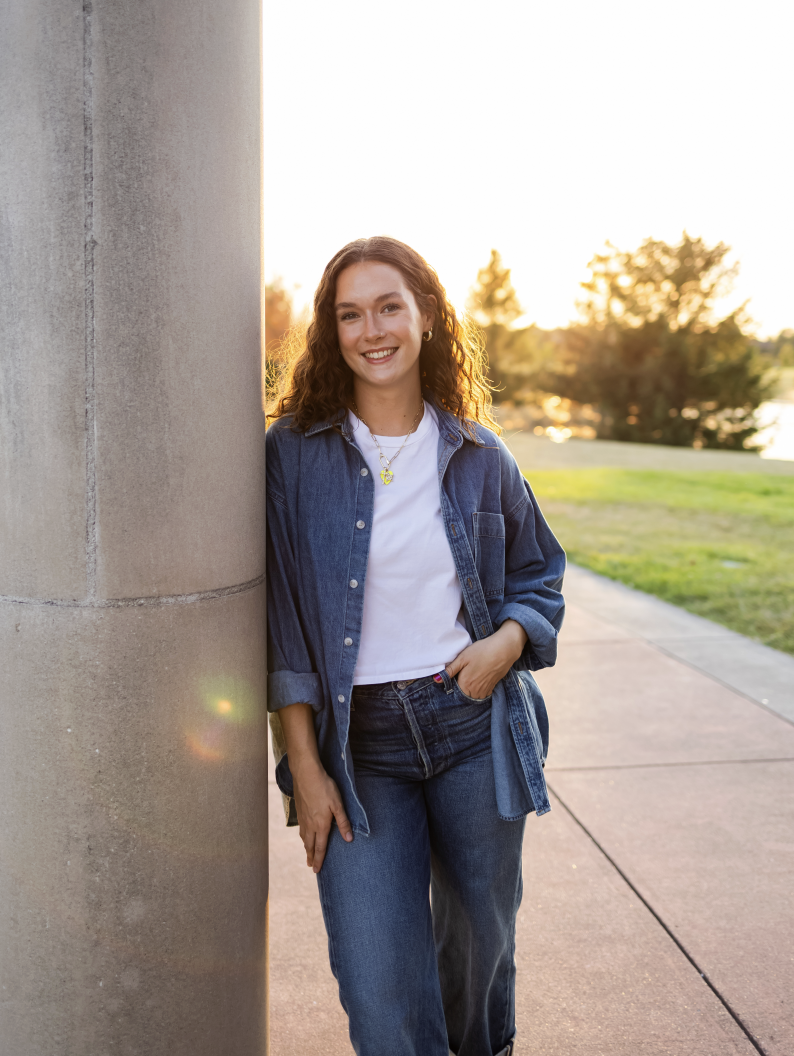 A young woman with curly brown hair smiling and leaning against a concrete pillar outdoors during sunset, wearing a denim jacket and jeans.