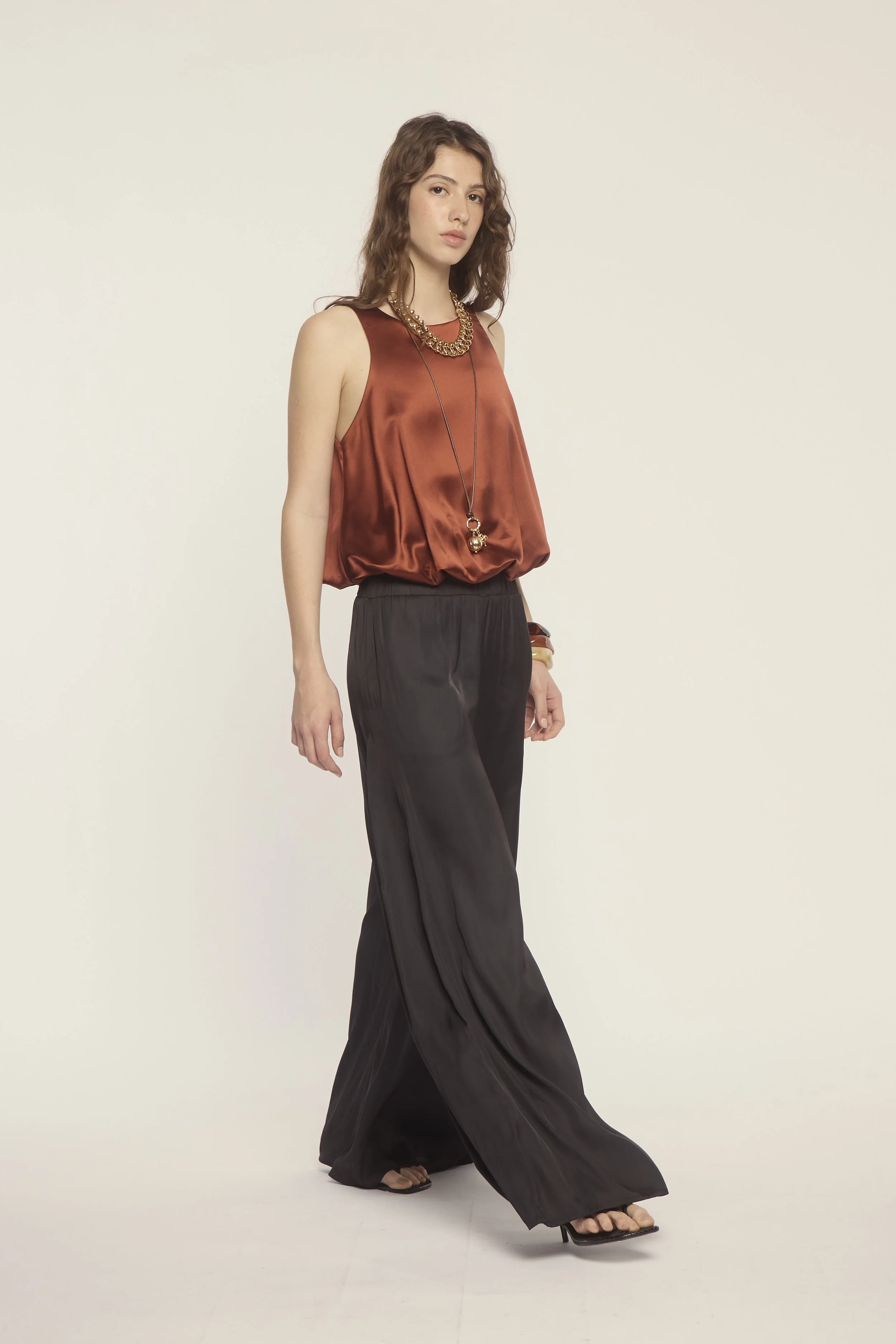 A woman in a rust-colored sleeveless satin top with black wide-leg pants, standing against a plain light-colored background.
