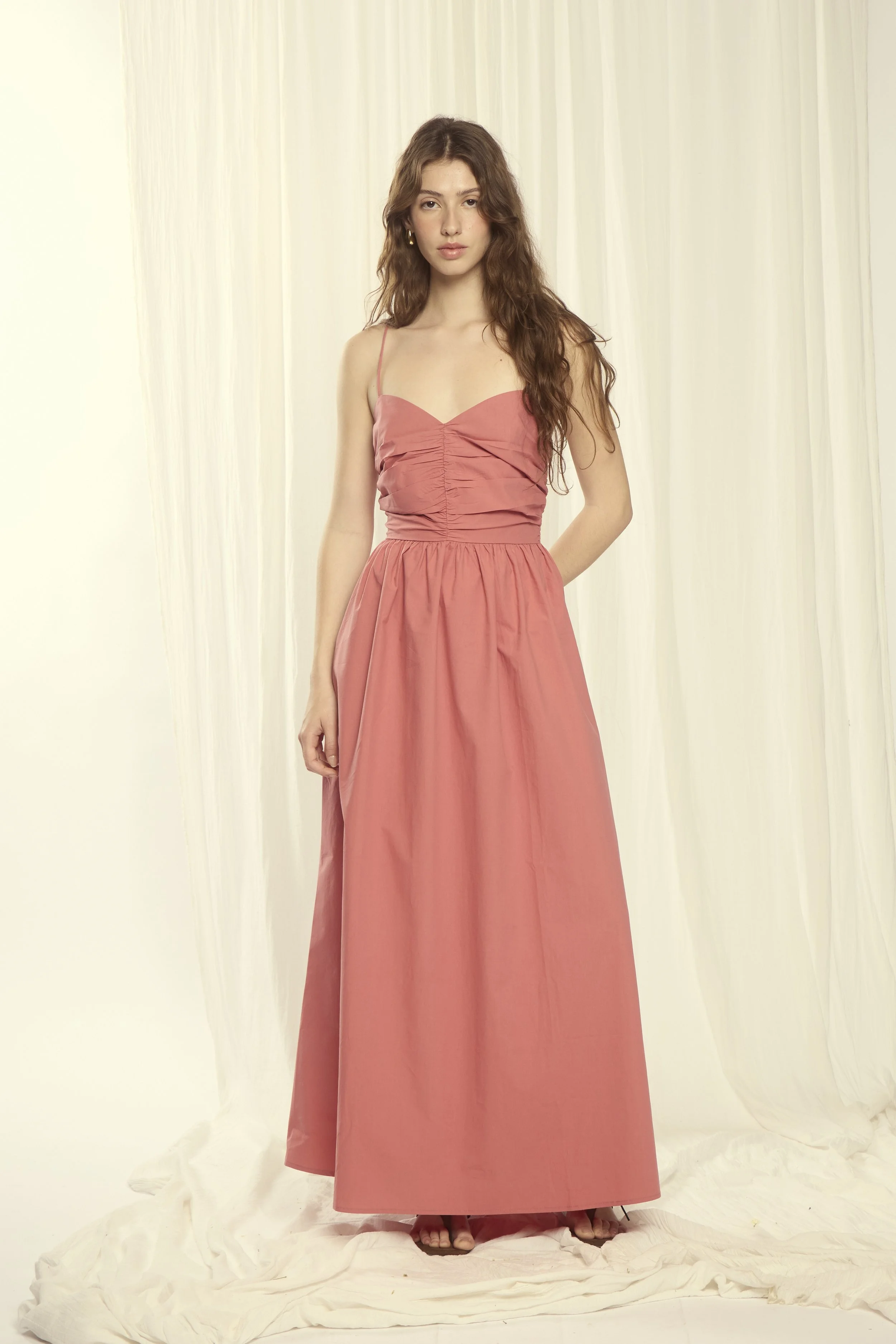 Young woman in a pink maxi dress standing on white fabric with a light-colored curtain background.