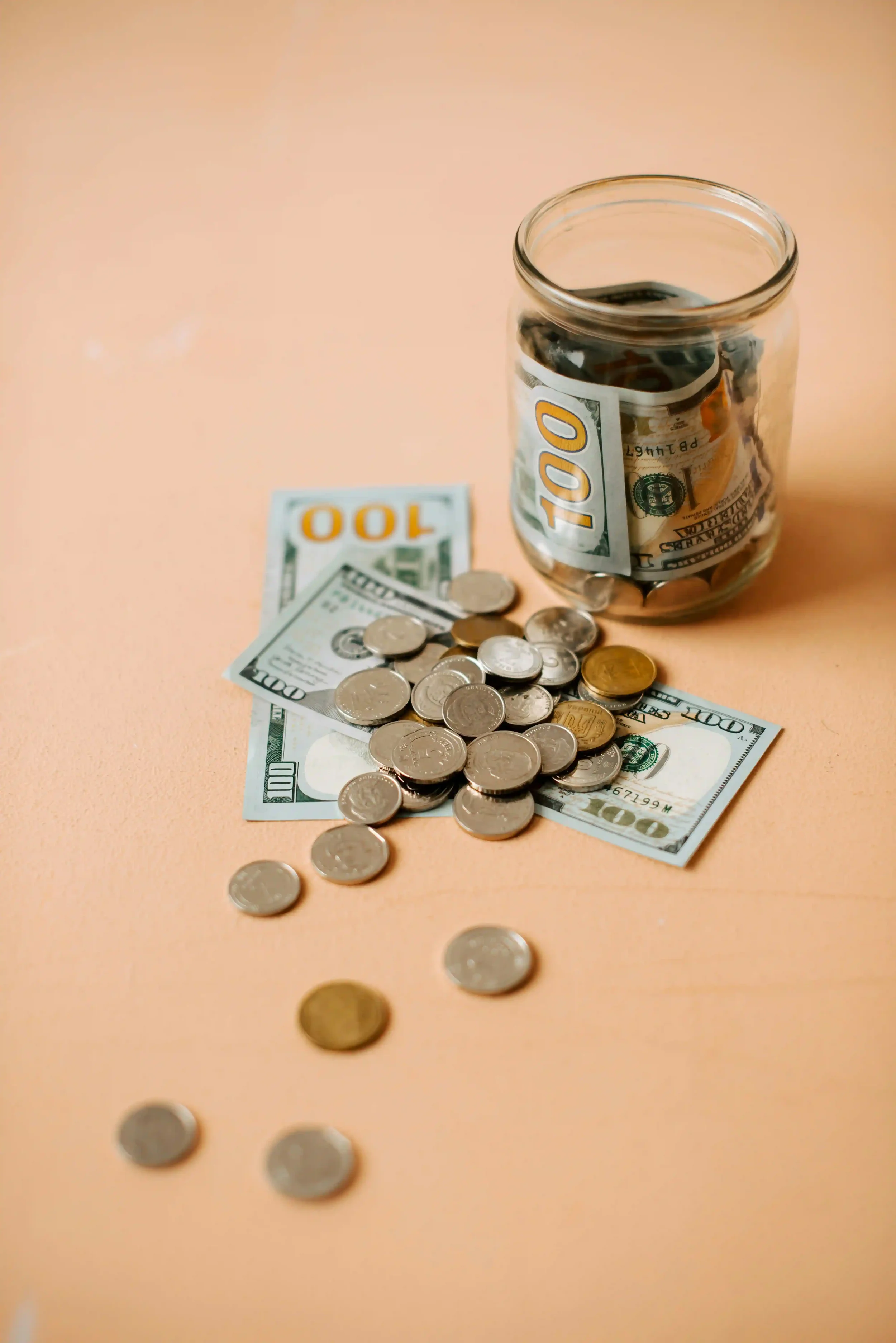 A glass jar filled with crumpled one hundred dollar bills and coins, resting on a light-colored surface, with additional bills and coins scattered around.