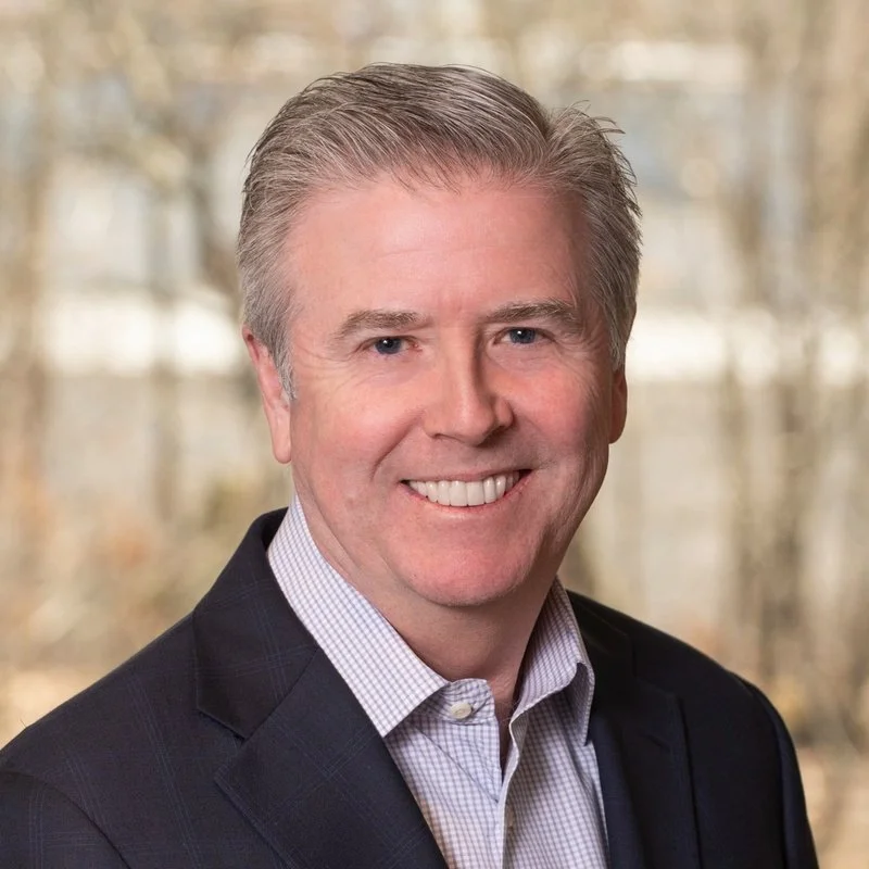 A smiling middle-aged man with light gray hair, wearing a dark blazer and a light checkered shirt, outdoors with trees in the background.