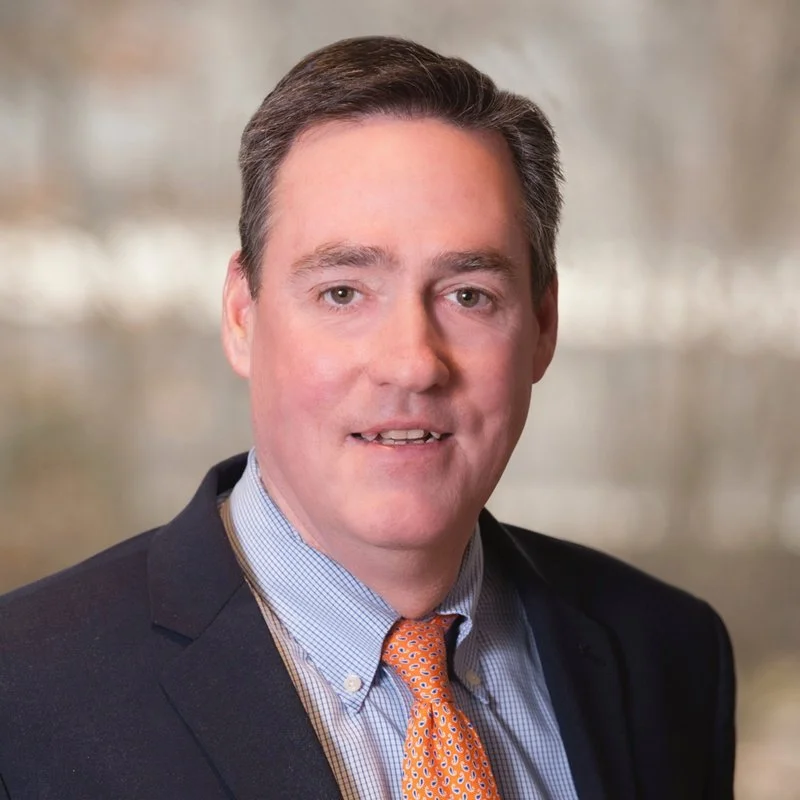 A man with short, dark hair, wearing a dark blazer, light blue dress shirt, and an orange patterned tie, looking at the camera with a neutral expression, standing outdoors with a blurred background.