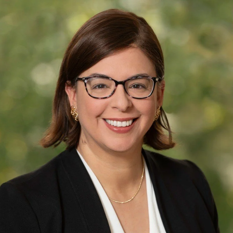 A woman with short brown hair, glasses, and gold jewelry smiling in front of a blurred green outdoor background.