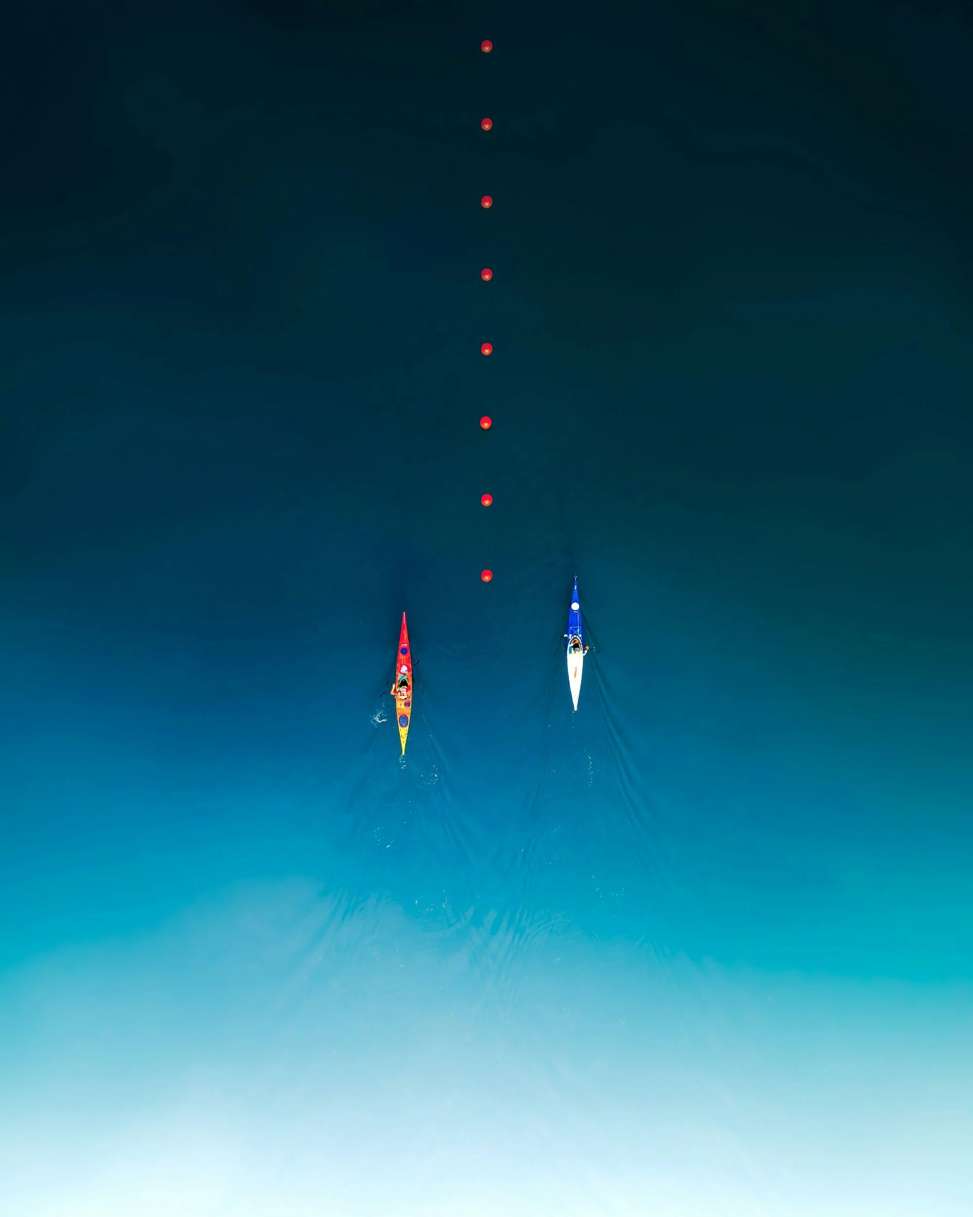 Two kayakers, one in a red kayak and one in a blue kayak, paddling in a calm body of water with a series of red buoys forming a line ahead.