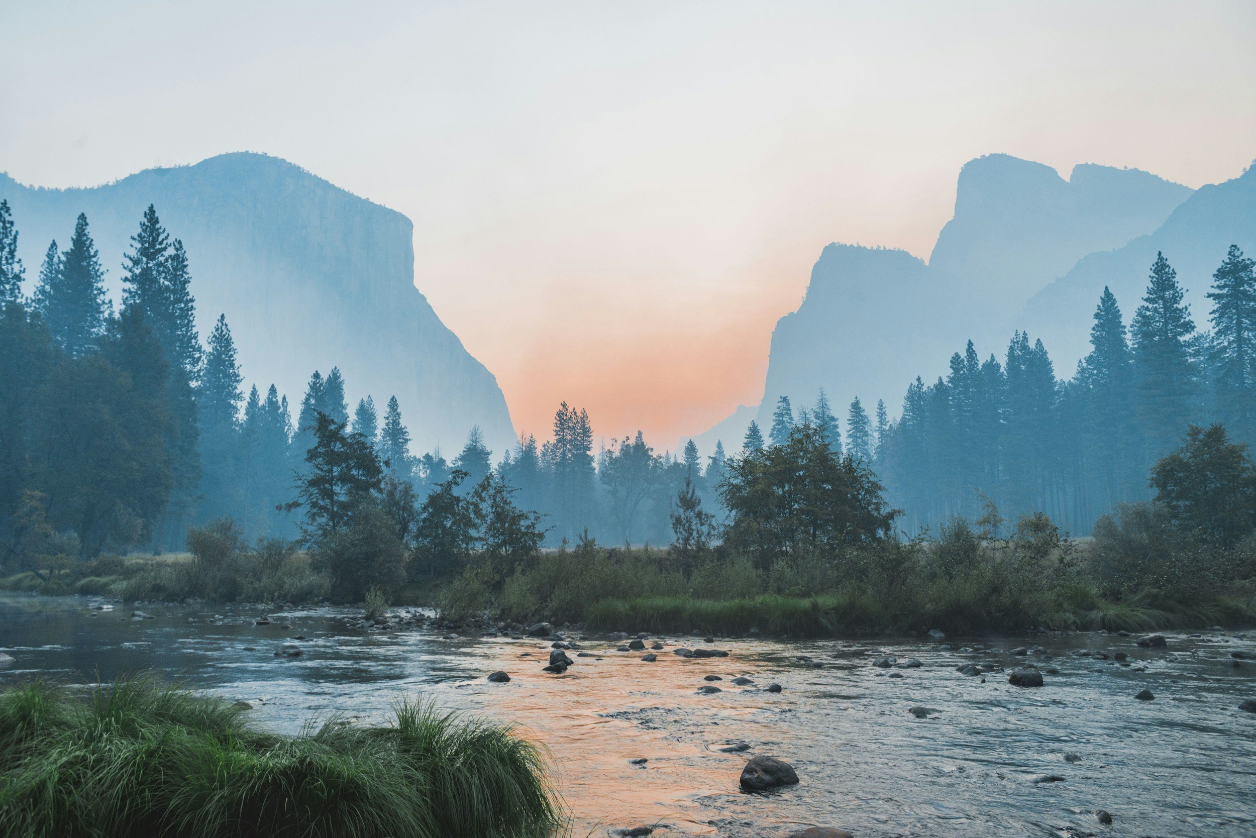 Scenic view of a river flowing through a forested valley with tall mountains in the background during dawn or dusk.