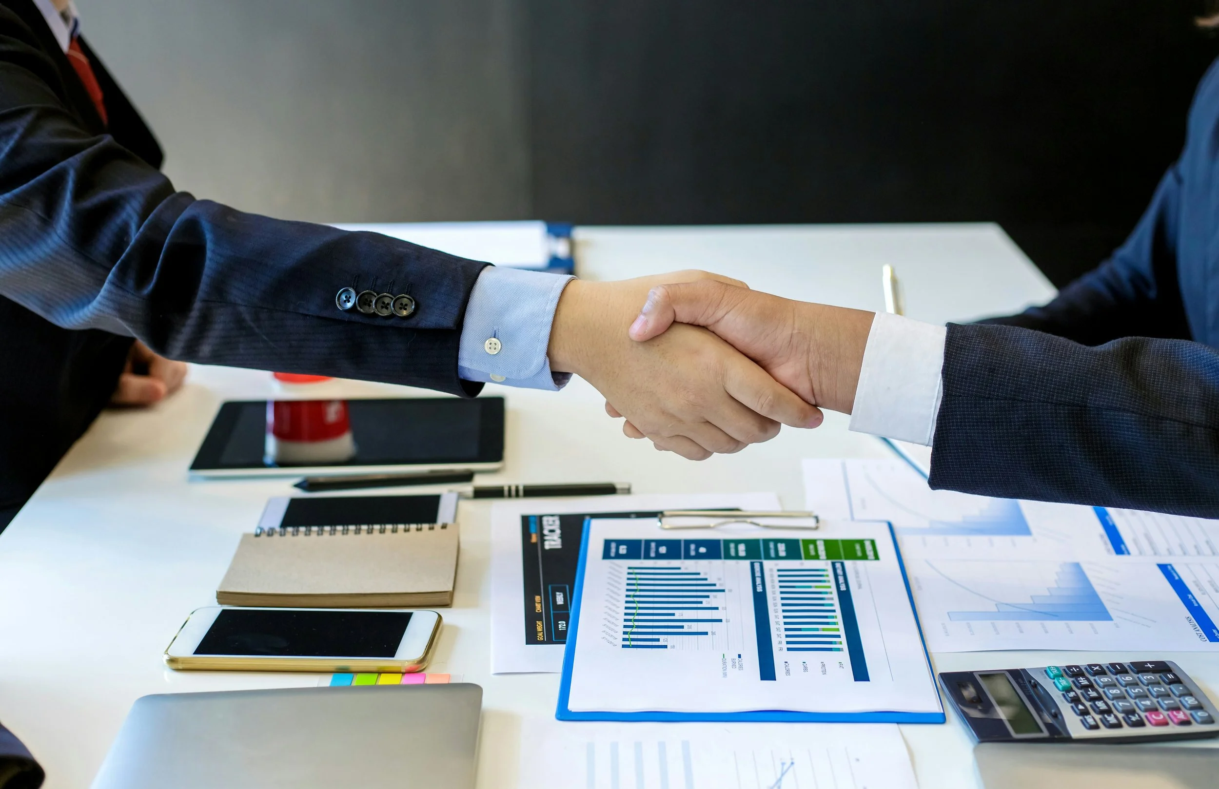 Two business people shaking hands over a conference table with documents, notebooks, a calculator, and electronic devices.
