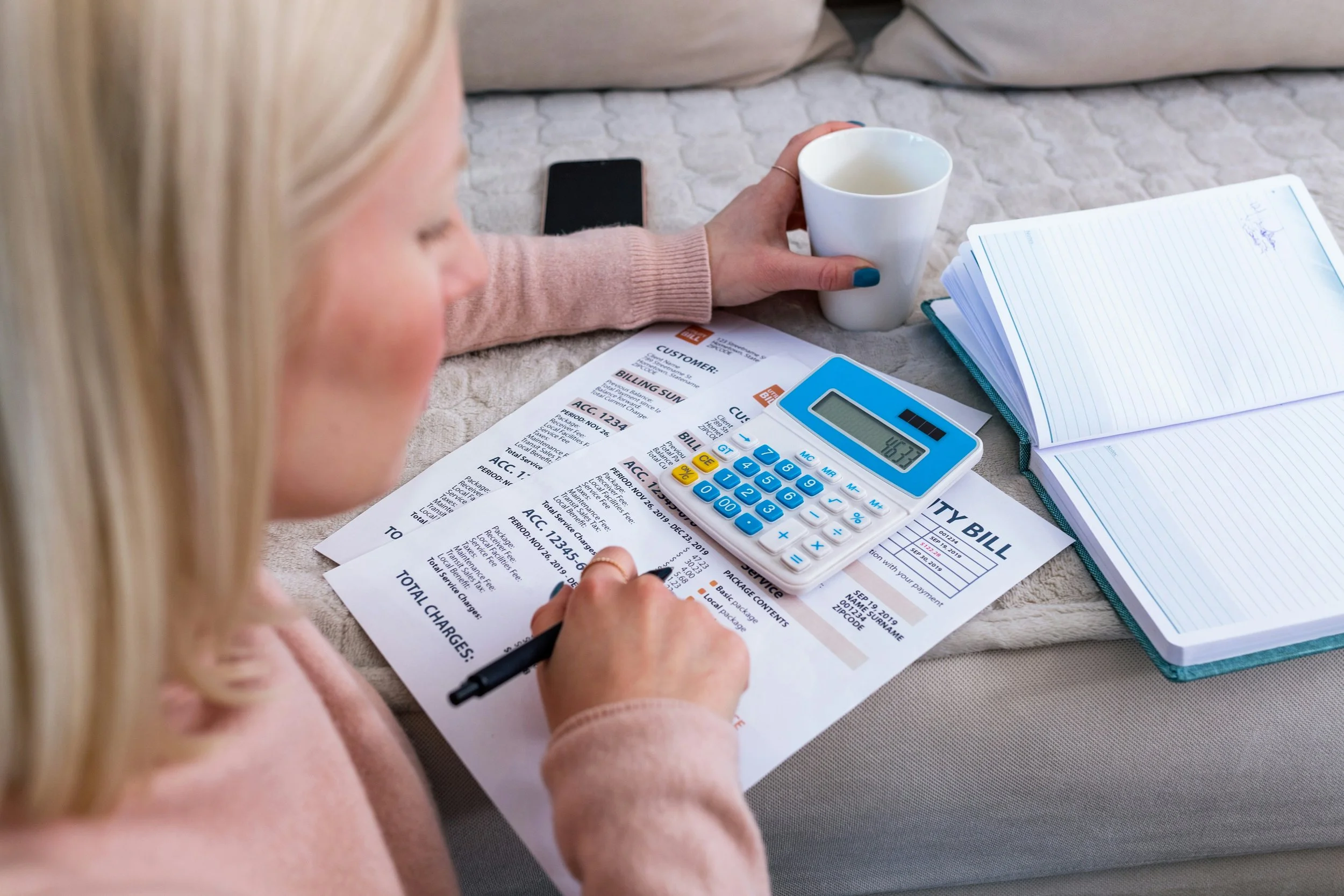 A woman filling out a utility bill on a bed, with a calculator, notebook, pen, mug, and smartphone on the bed.