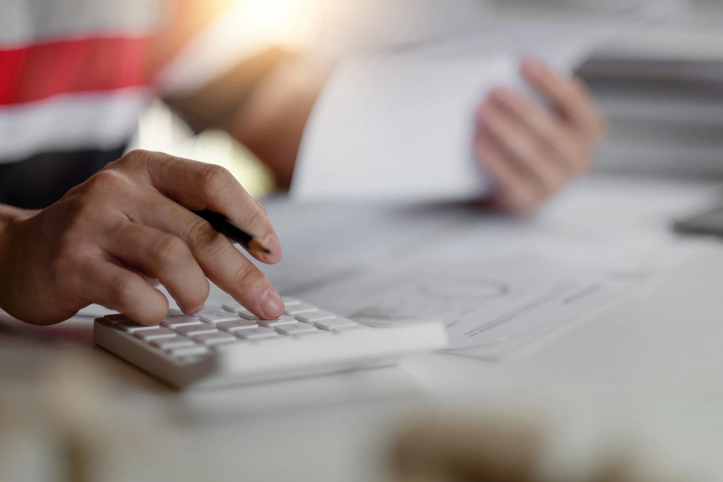 Close-up of a person's hand pressing keys on a white calculator, with a pen resting on the person's hand. In the background, a person is holding a white tablet or notebook, slightly out of focus.