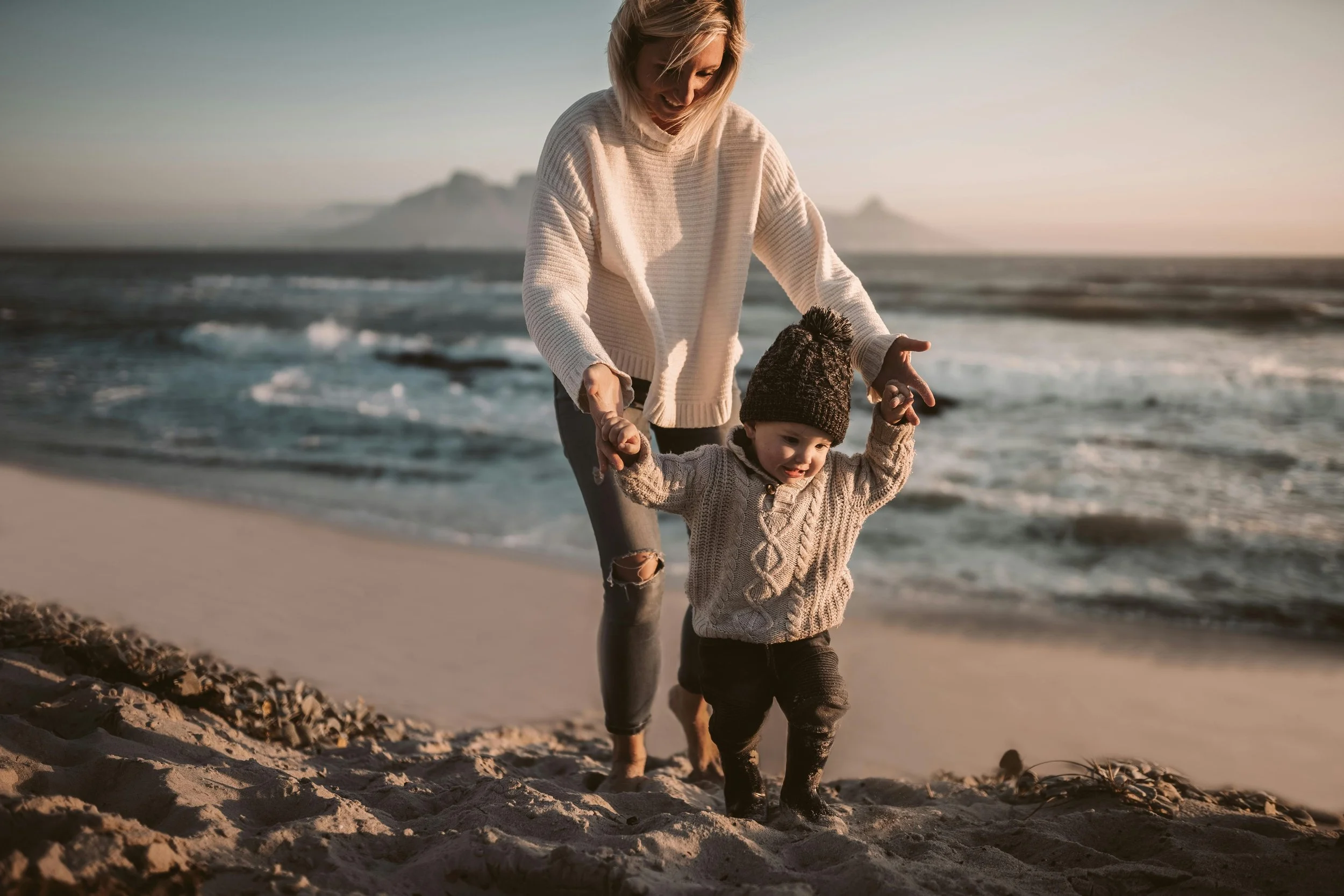 A woman and a young child walking on a sandy beach near the ocean during sunset, holding hands and smiling.