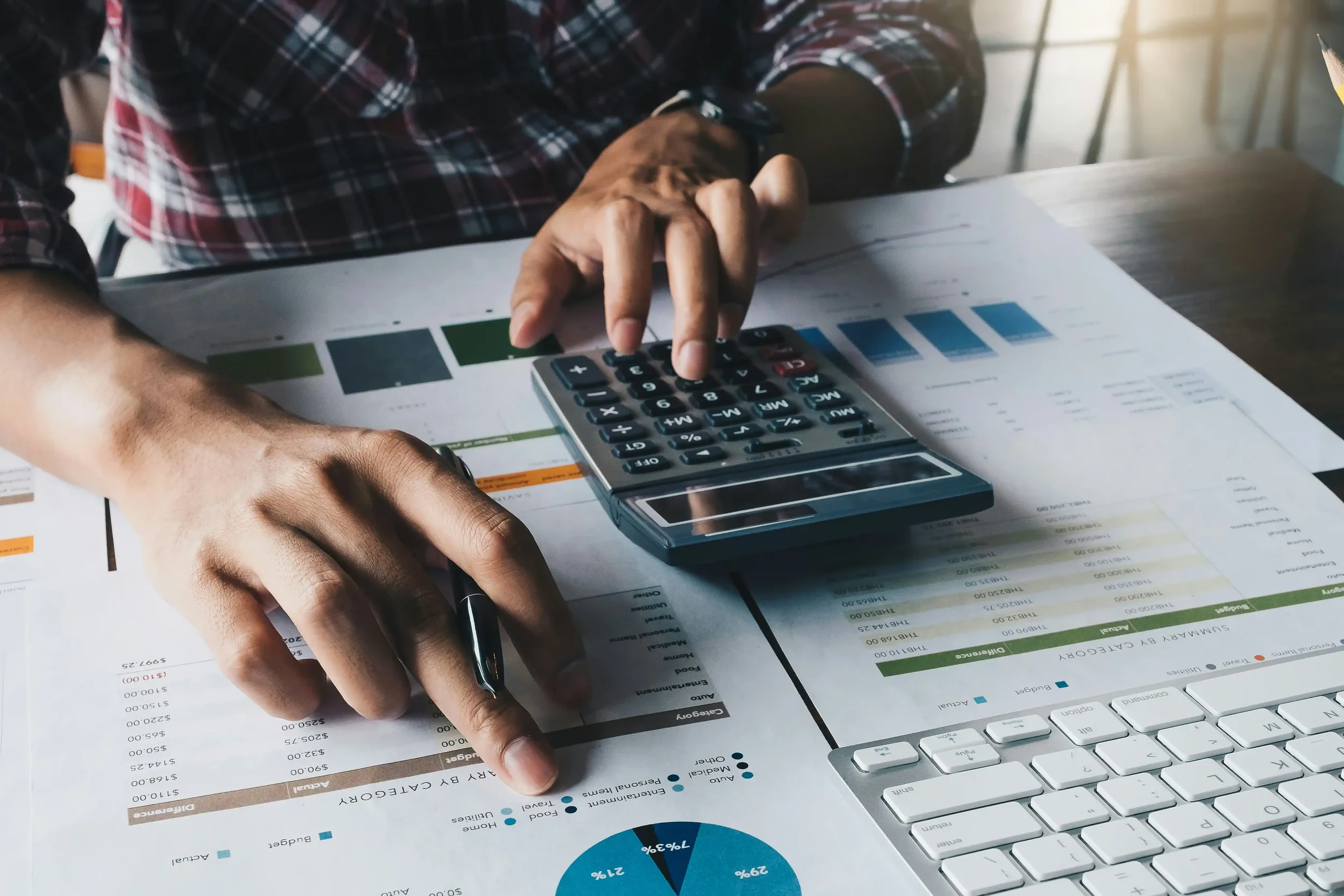 Person using a calculator and a pen, working on financial documents and charts on a desk, with a keyboard nearby.