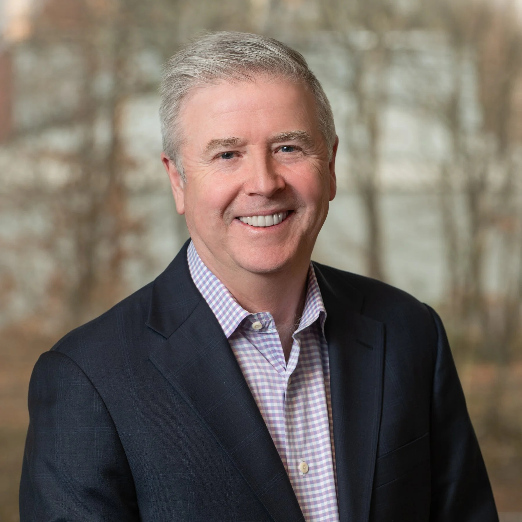 A smiling middle-aged man with light gray hair, wearing a dark blazer and a light checkered shirt, outdoors with trees in the background.