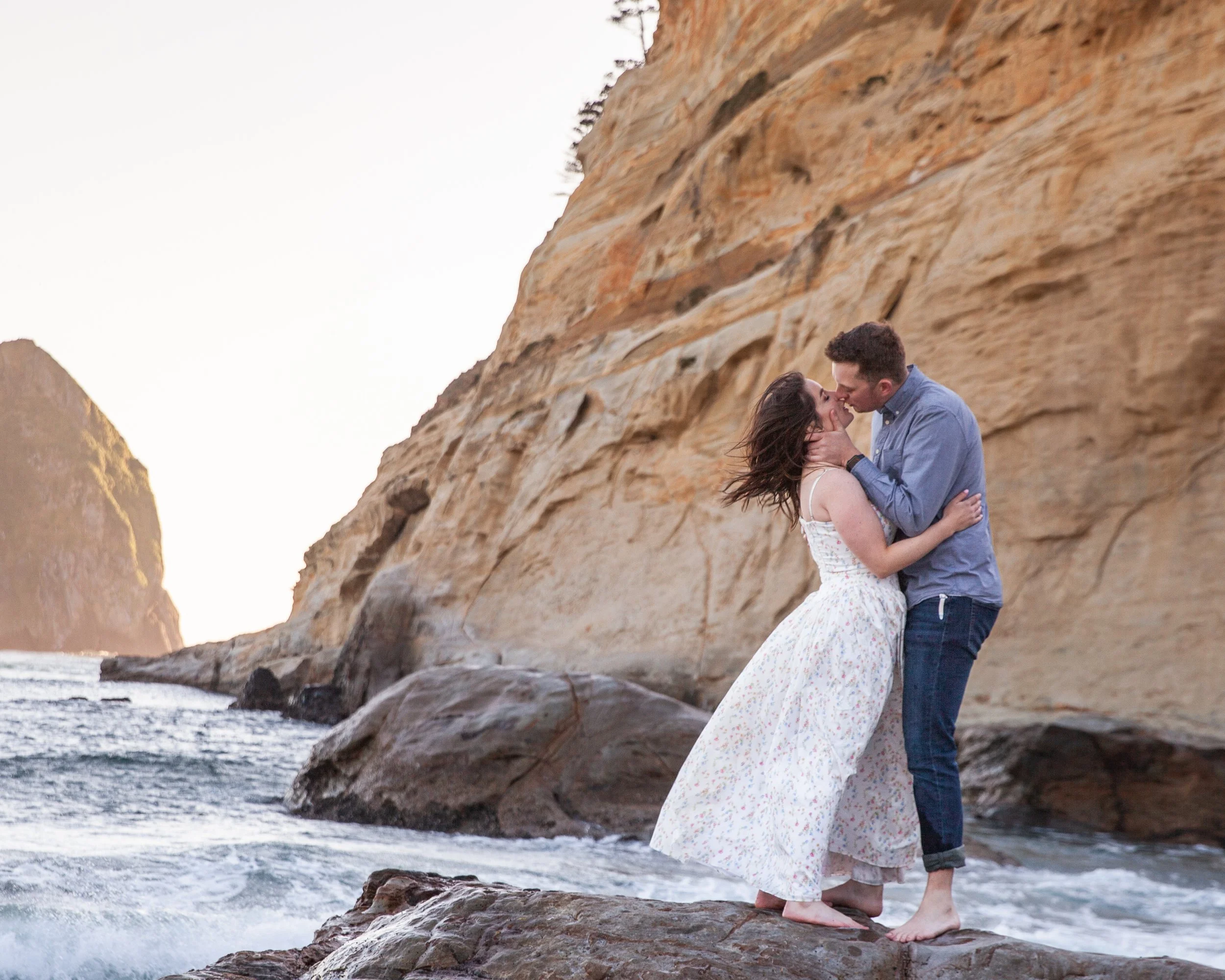 Couple embracing beneath dramatic coastal cliffs during an editorial engagement session. Oregon Coast.