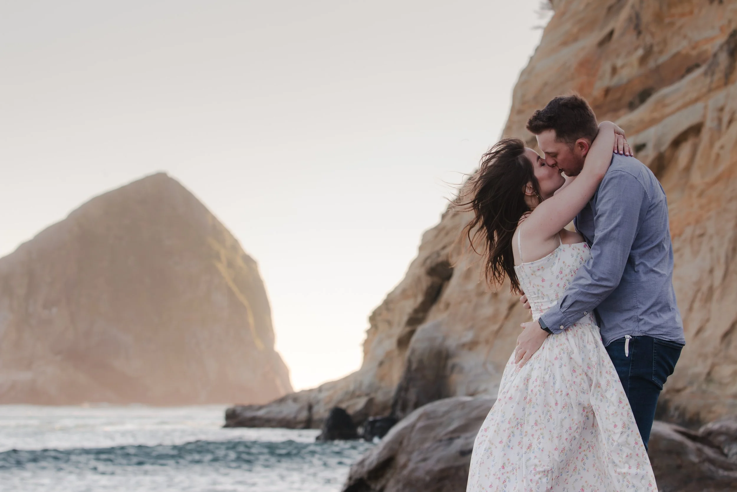 Couple embracing beneath dramatic coastal cliffs during an editorial engagement session. Oregon Coast.