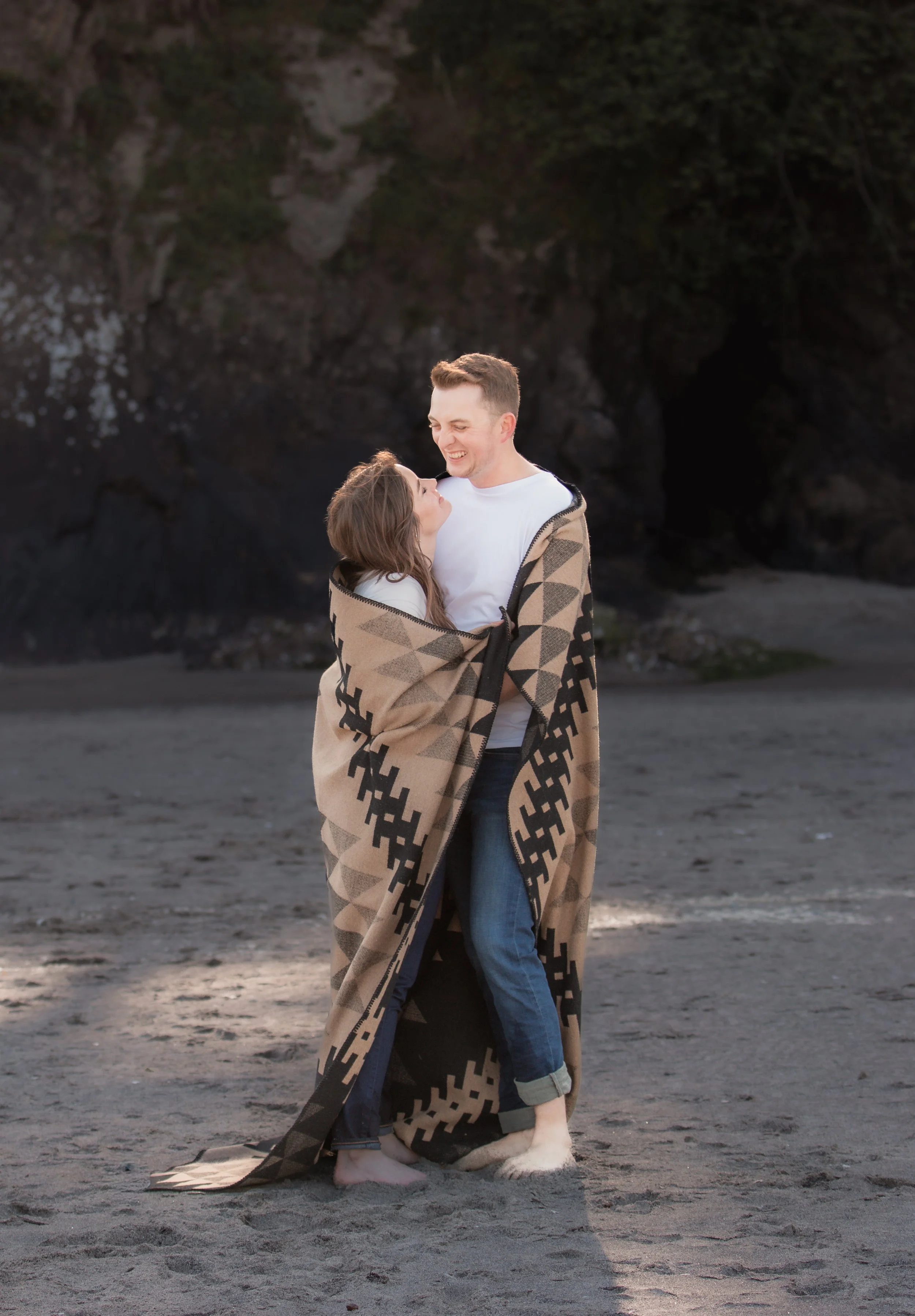 Couple embracing beneath dramatic coastal cliffs during an editorial engagement session. Oregon Coast. 
