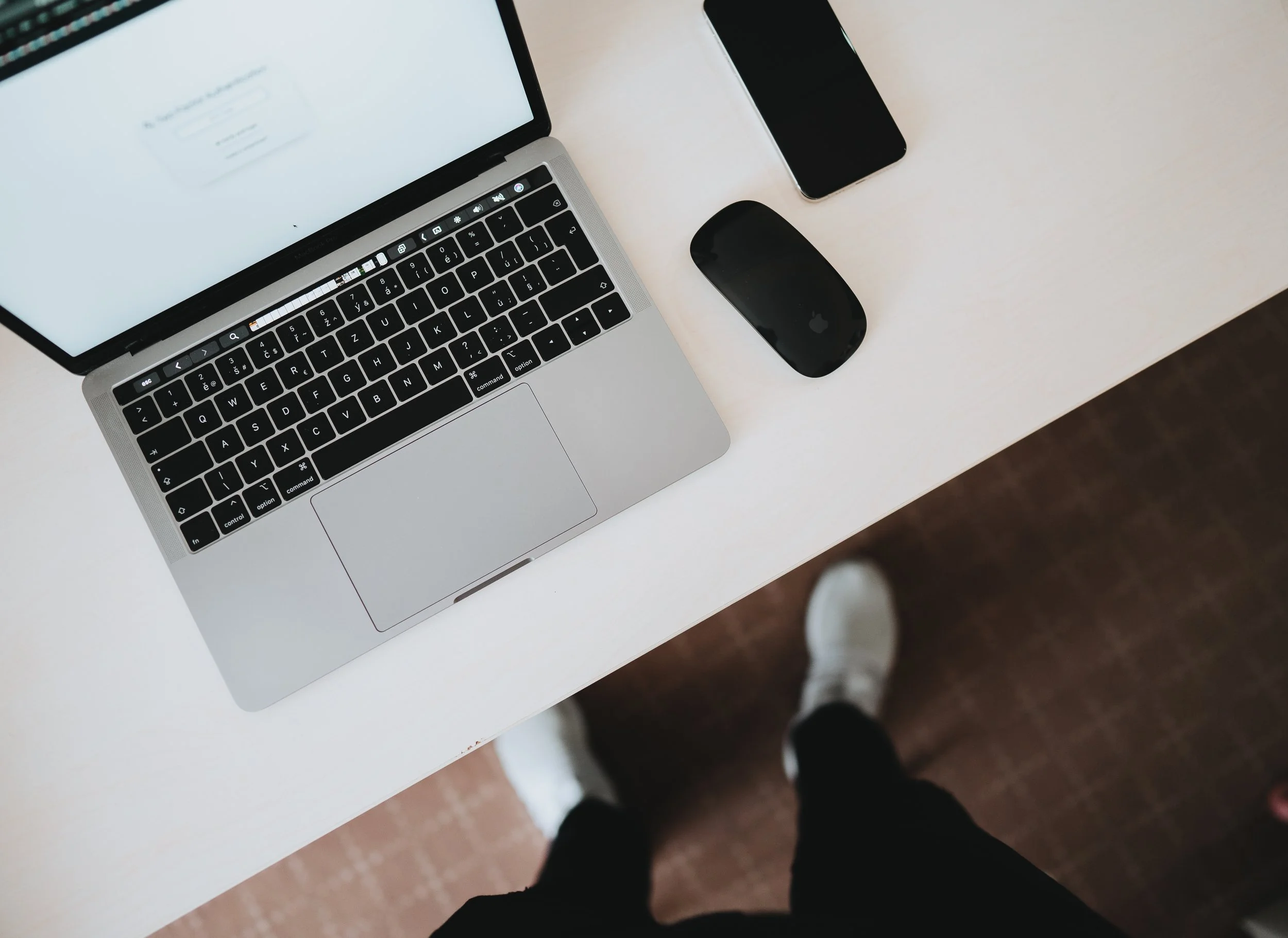 Top-down view of a workspace with a silver laptop, a black mouse, and a smartphone on a white desk. A person's legs and white sneakers are visible at the bottom of the image.