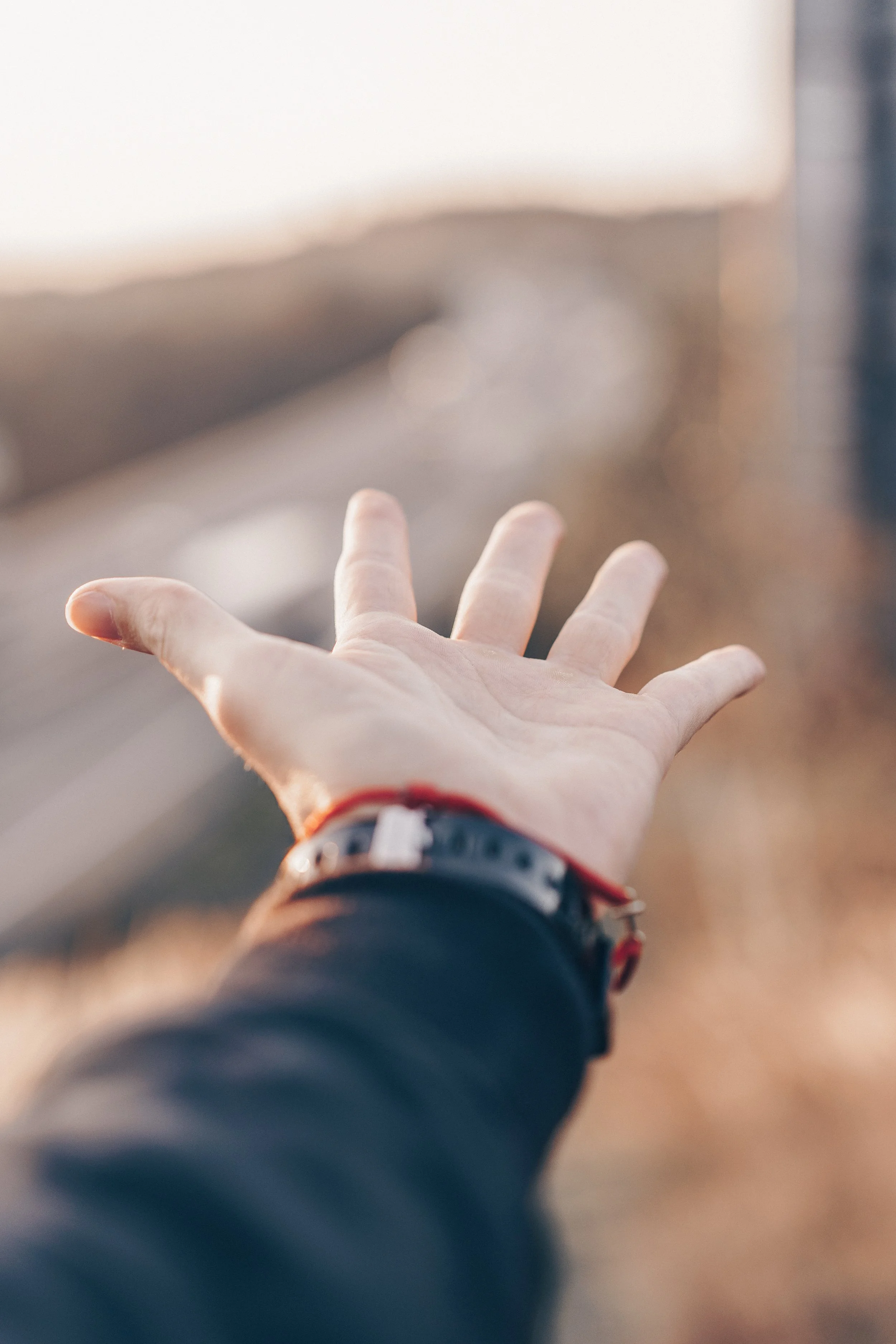 A person's outstretched hand reaching towards a blurry background outdoors, wearing a black watch with a red strap.