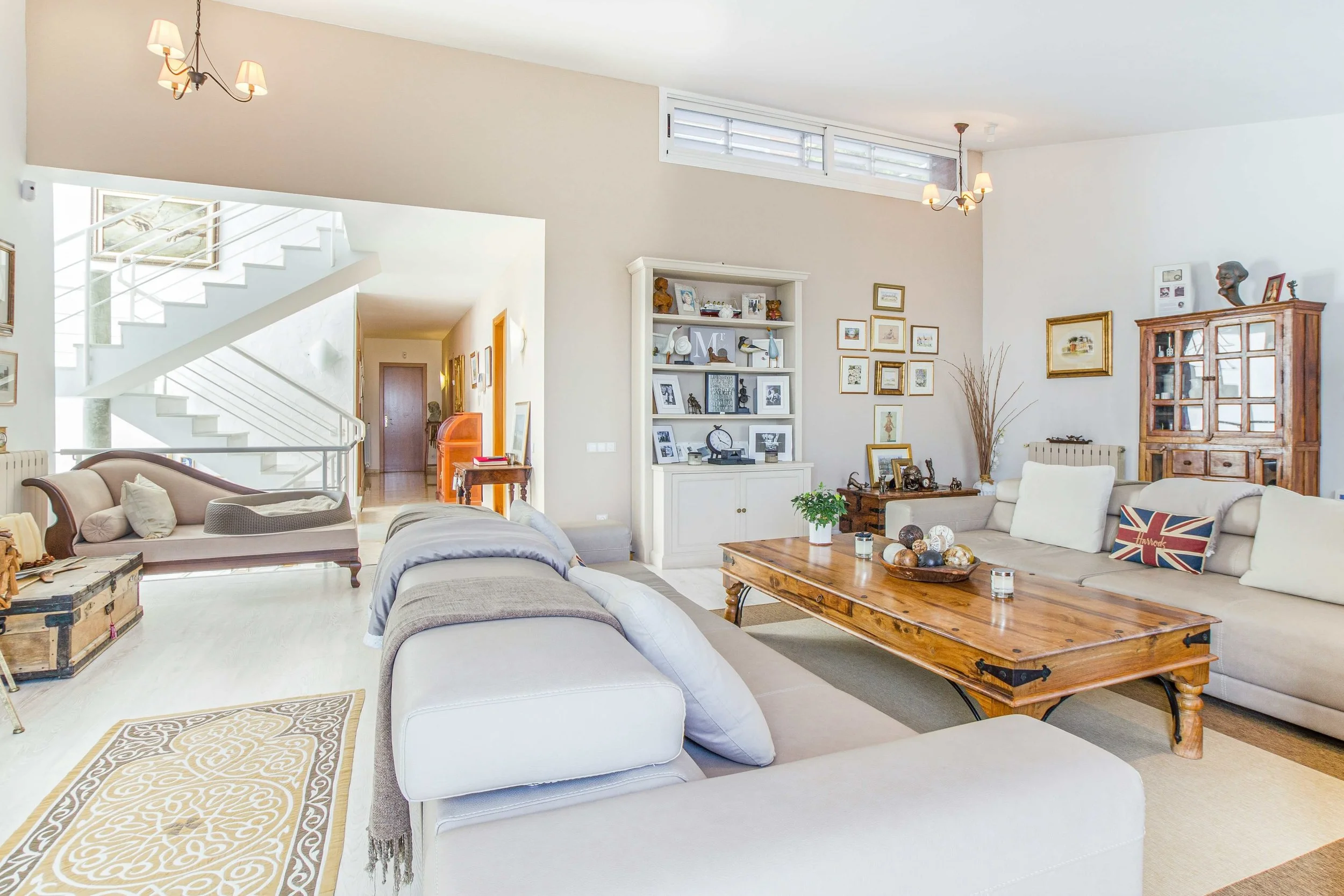 Bright living room with white sofas, a wooden coffee table, decorative pillows, and wall art. There is a white bookshelf with framed photos and ornaments, a wooden cabinet, and a staircase in the background.