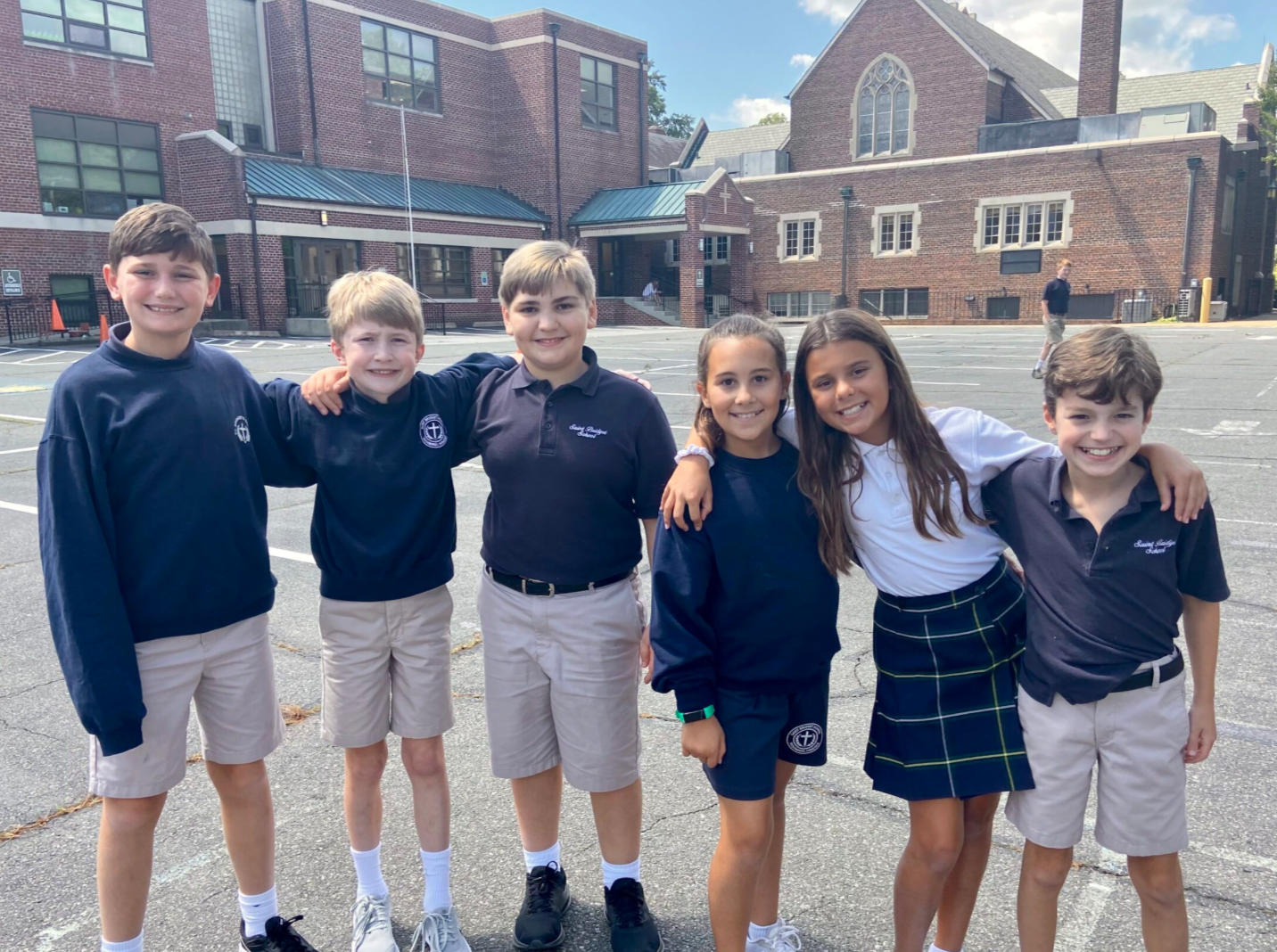 Six children standing together outdoors in a parking lot with school buildings in the background.