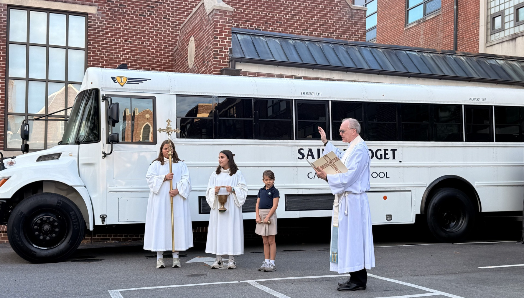A priest performing a religious ceremony outside, with three children dressed in white robes, in front of a white bus marked 'SAINT BRIDGET SCHOOL' in a city setting.