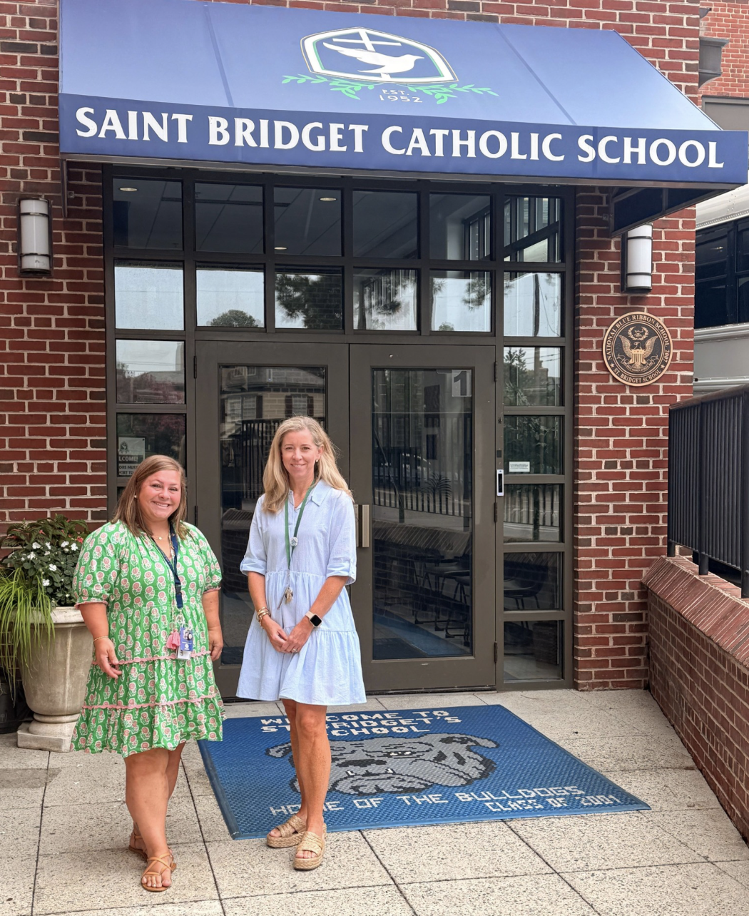 Principal and Vice Principal standing in front of the entrance to Saint Bridget Catholic School, with a blue awning and a school emblem above the door.