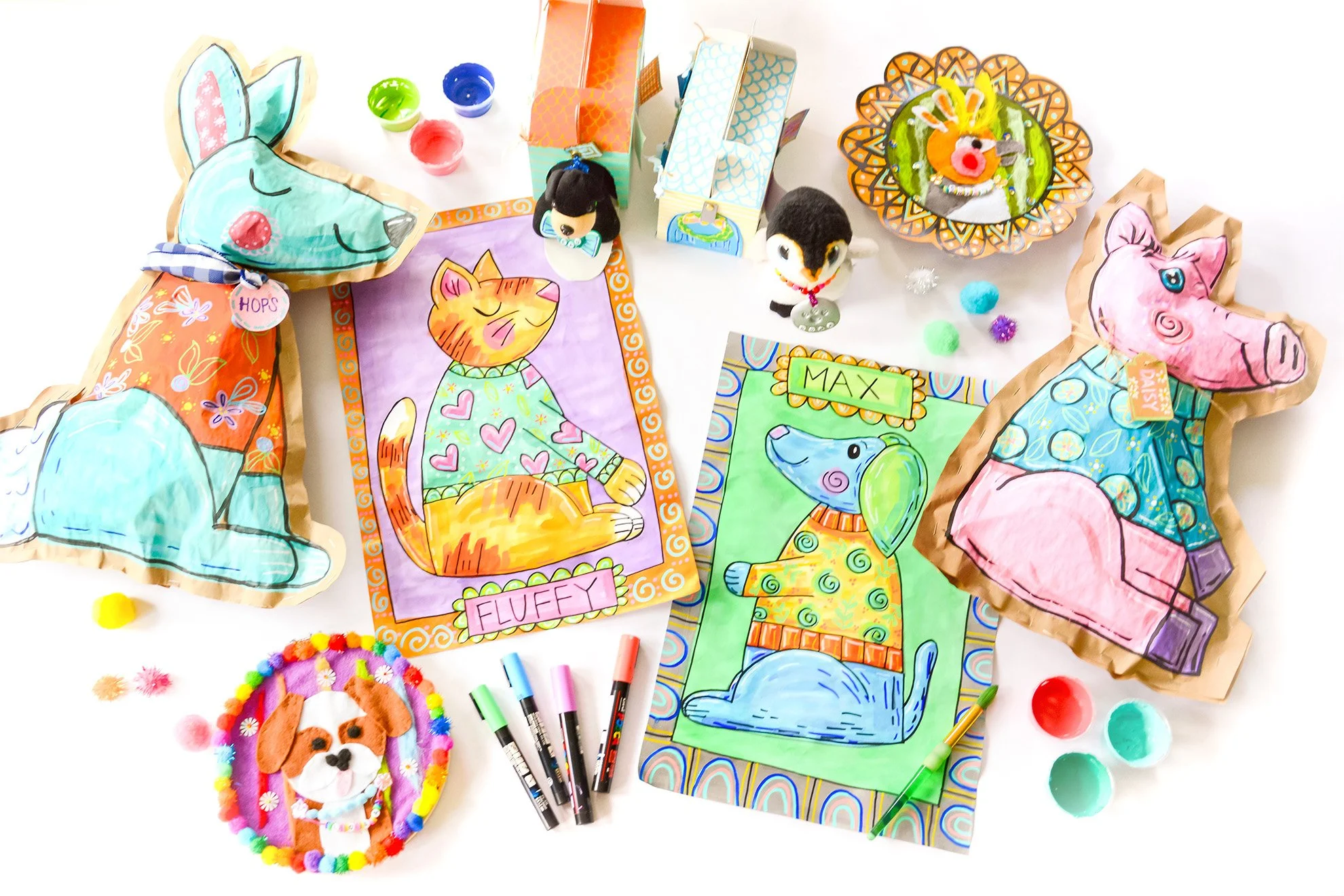 Colorful handmade animal-themed greeting cards and crafts on a white table, including a fox, pig, cat, and dog, with markers, pom-poms, and paint
