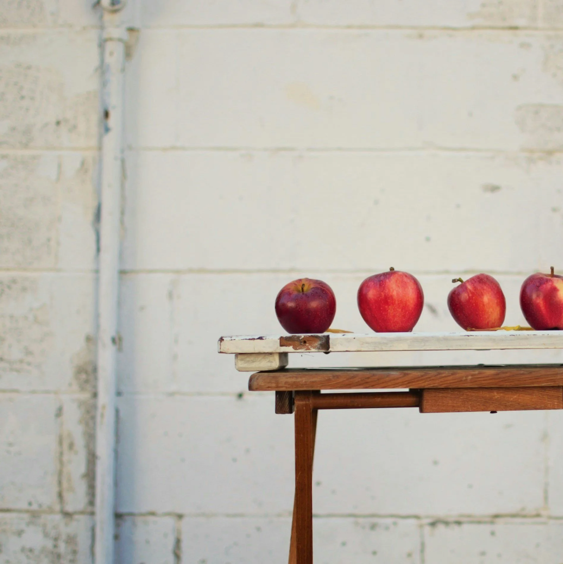 Four red apples on a wooden table against a white painted brick wall.