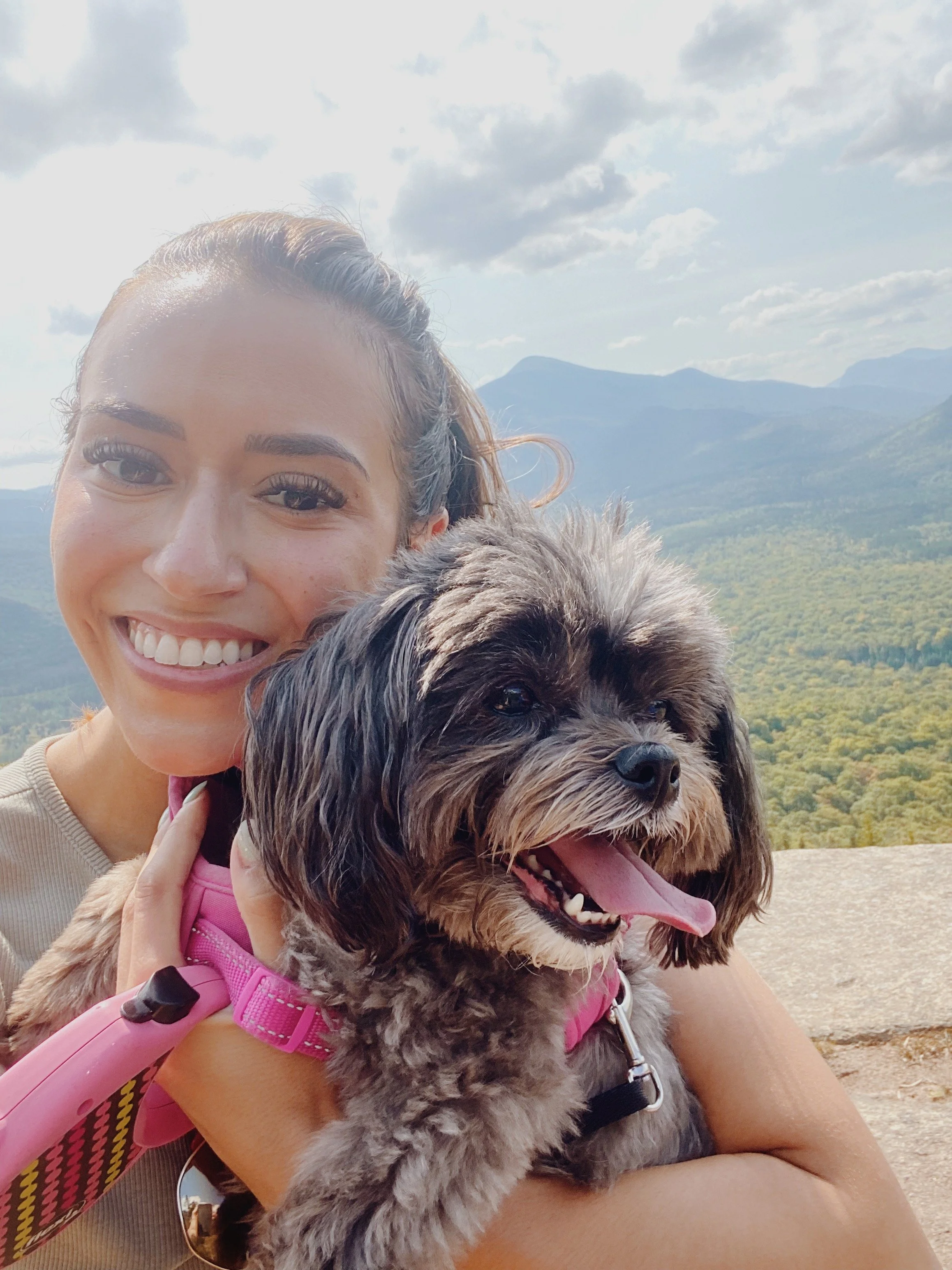 A woman smiling and holding a fluffy dog outdoors with mountains and cloudy sky in the background.