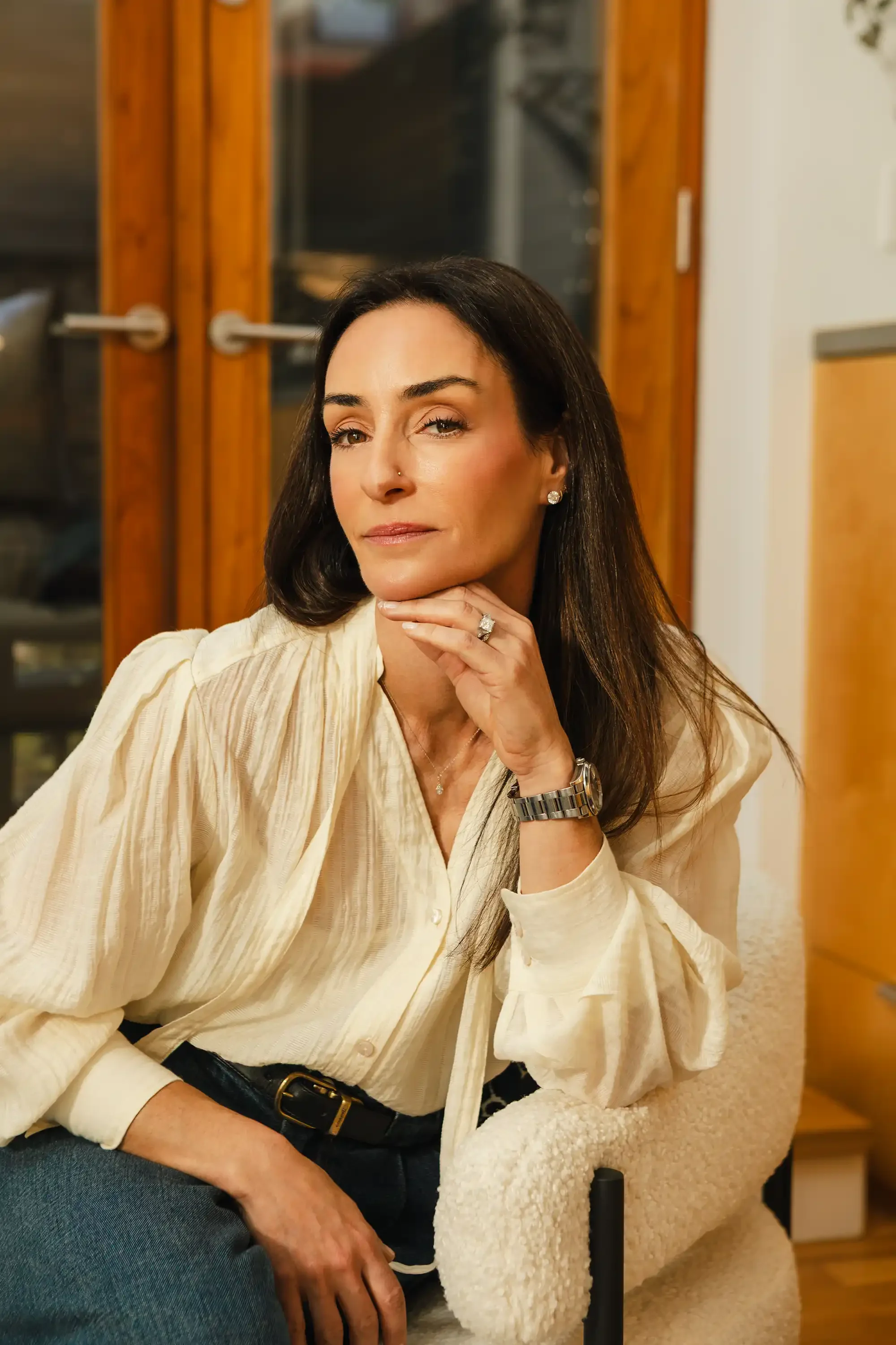 A woman with dark hair and a nose piercing sitting on a beige armchair, wearing a cream-colored blouse, a gold watch, earrings, a ring, and a necklace, inside a room with wooden doors and a drawer in the background.