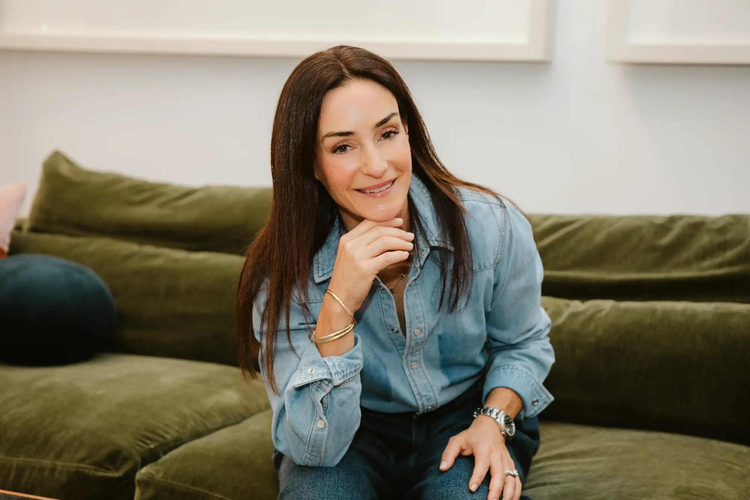 A woman with long brown hair, smiling, sitting on a green couch, resting her chin on her hand.