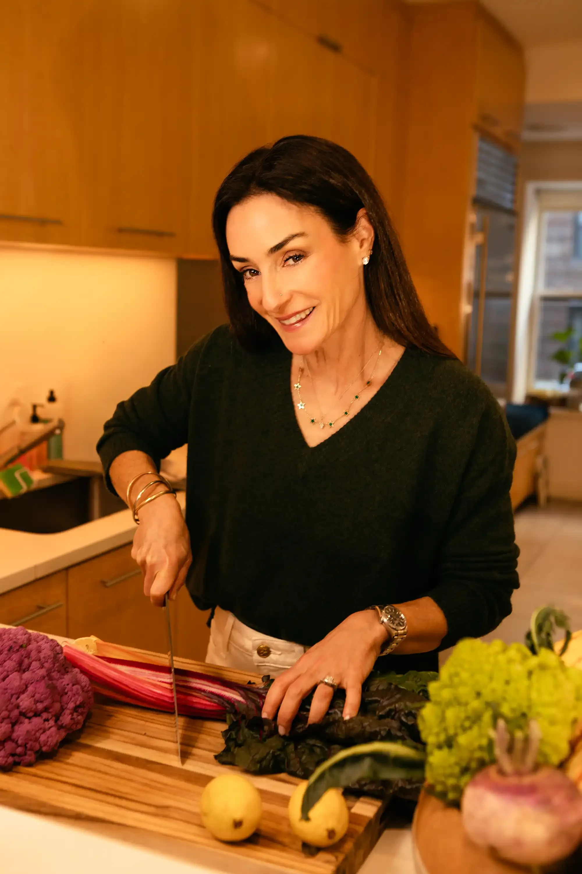 A woman with dark hair and jewelry is chopping vegetables on a kitchen countertop, surrounded by colorful produce.