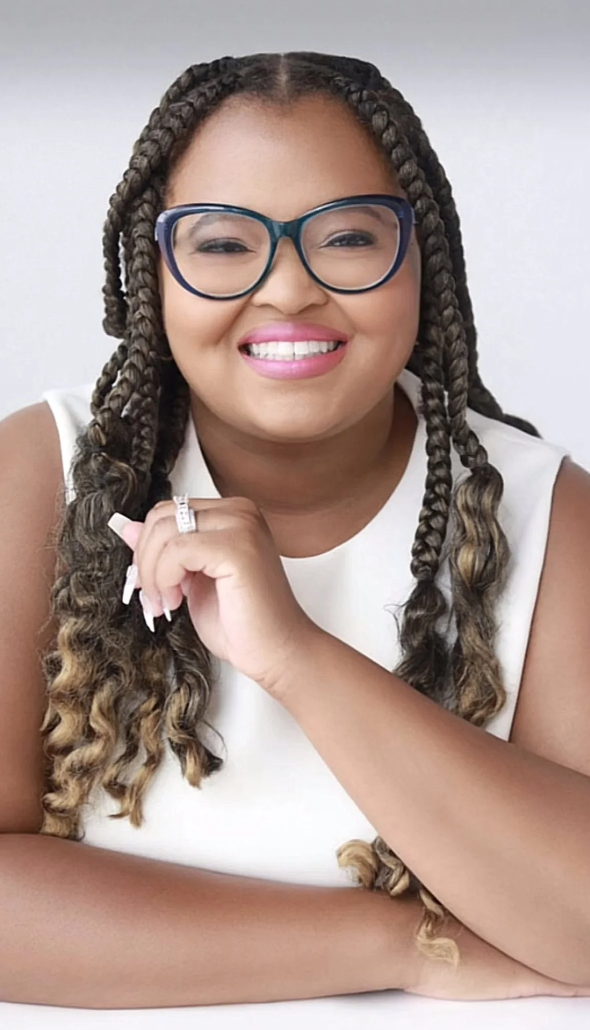 A smiling woman with long braided hair, wearing glasses and a white sleeveless top, posing against a plain background.