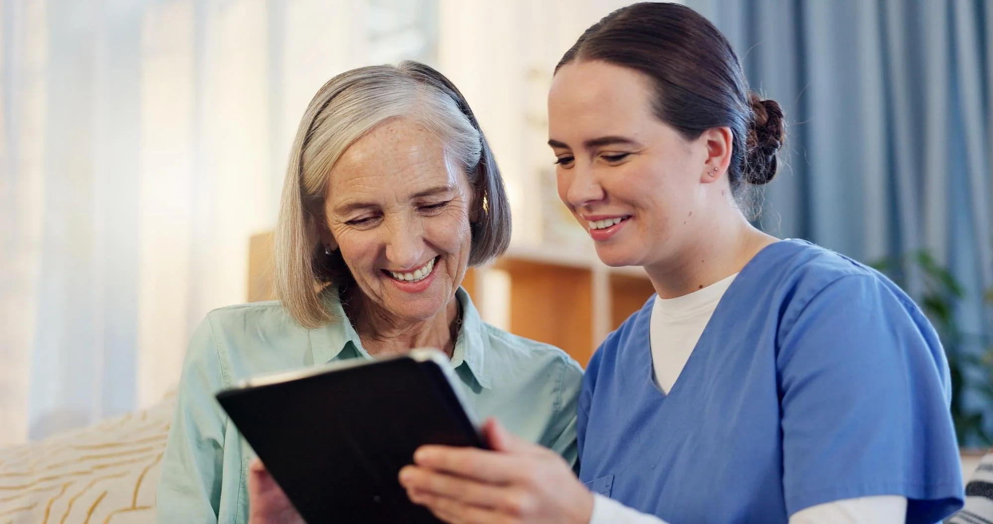 A senior woman and a young female healthcare worker smiling while looking at a tablet together indoors.