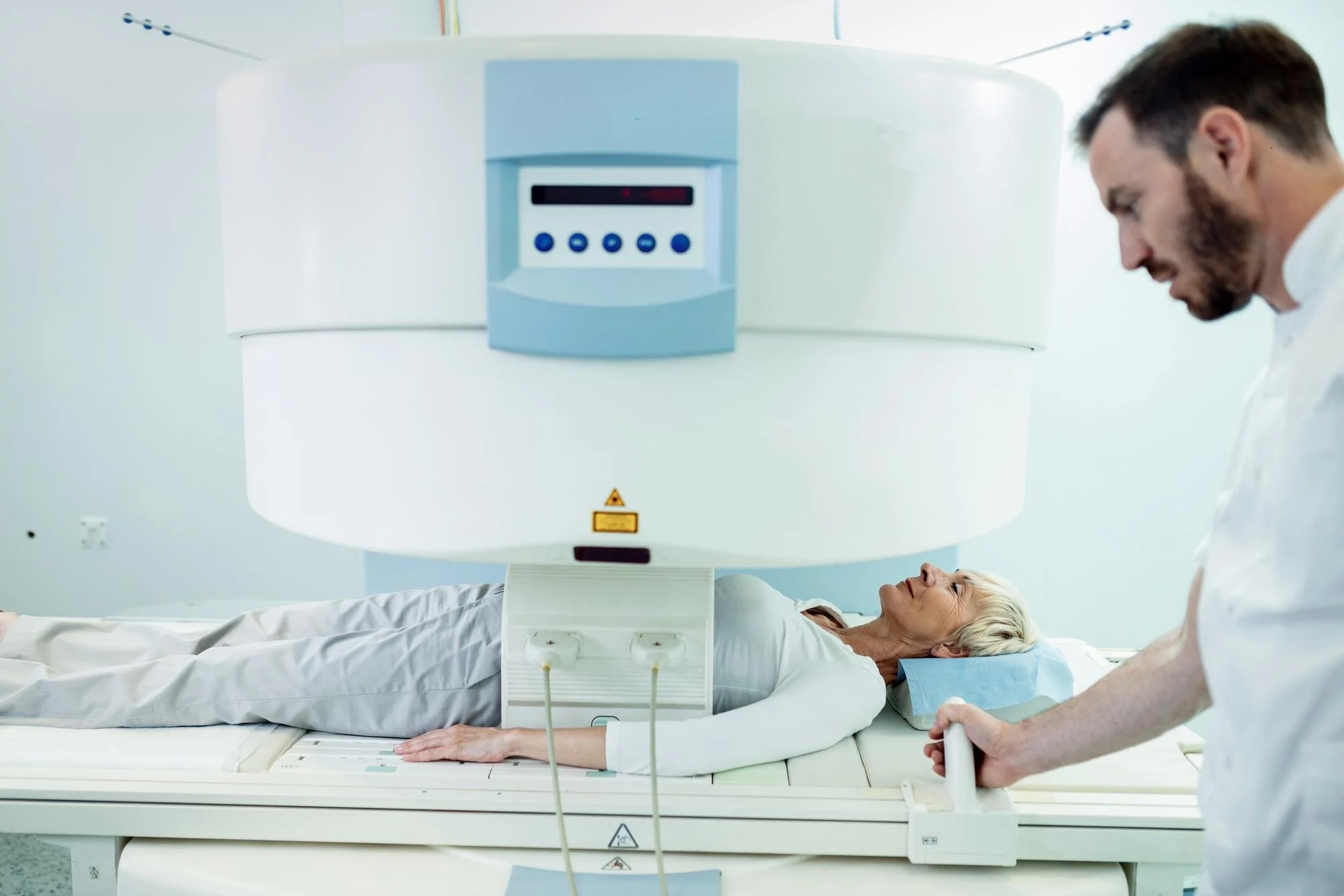 A woman lying on a medical scanner table while a technician or doctor prepares her for an MRI scan in a clinical setting.