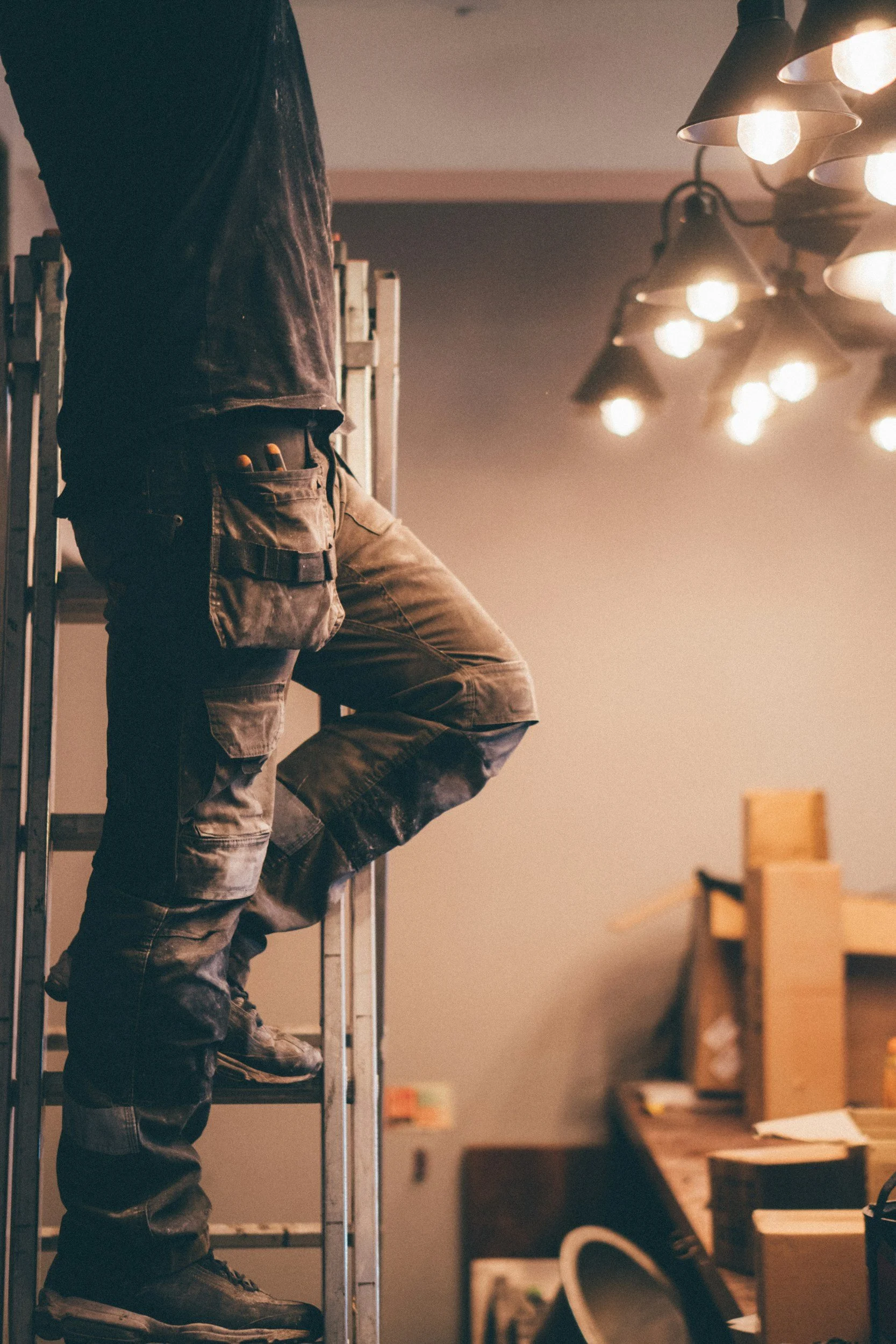A person standing on a ladder in a workshop or industrial space, wearing work pants and a tool pouch, with a cluttered workspace in the background and multiple light fixtures overhead.